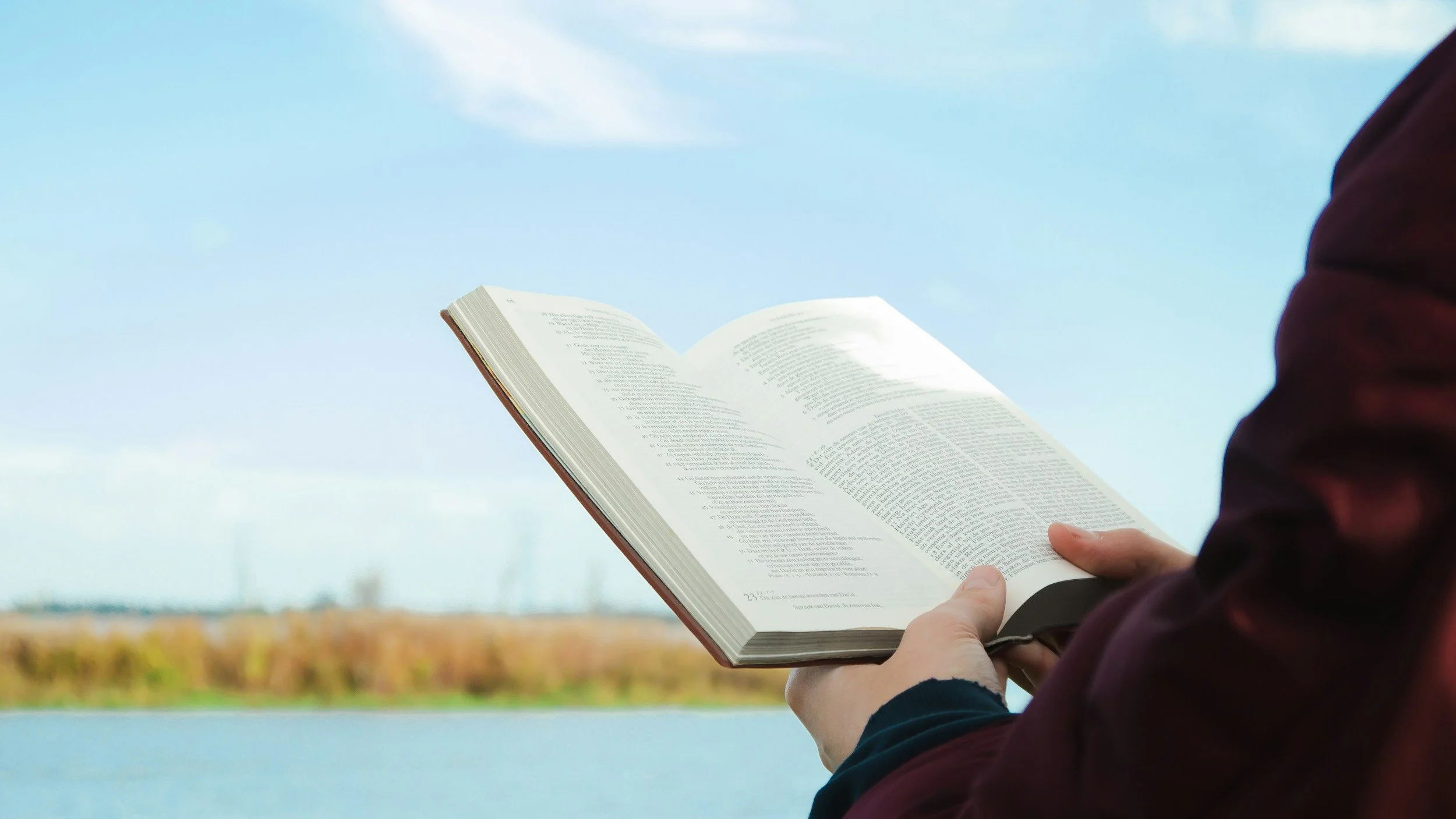 A person in a burgundy jacket holding an open book outdoors with a body of water and a cloudy sky in the background.