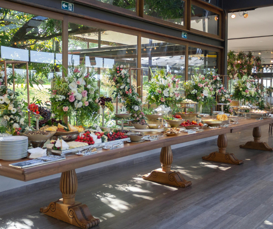 Buffet table with various dishes and large floral arrangements inside a well-lit restaurant with large windows.