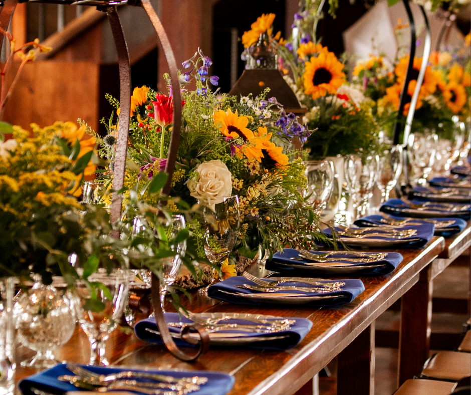A long wooden table set for a celebration with floral centerpieces, navy blue napkins, and silverware, in a rustic indoor setting.