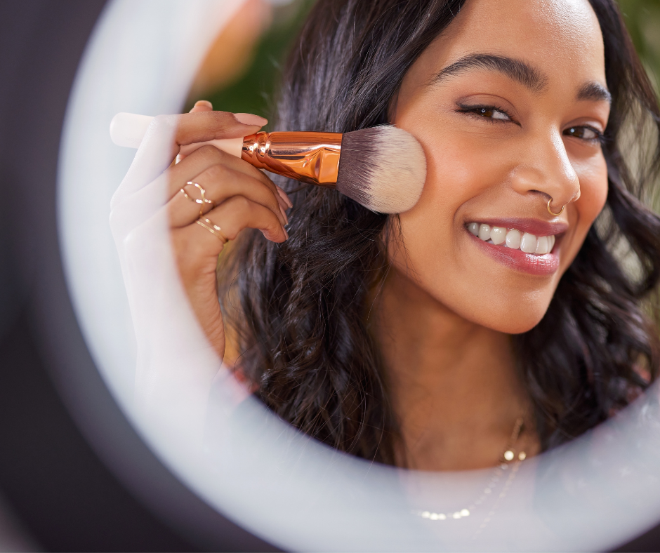 A woman with dark hair and a nose ring smiling while applying makeup with a brush