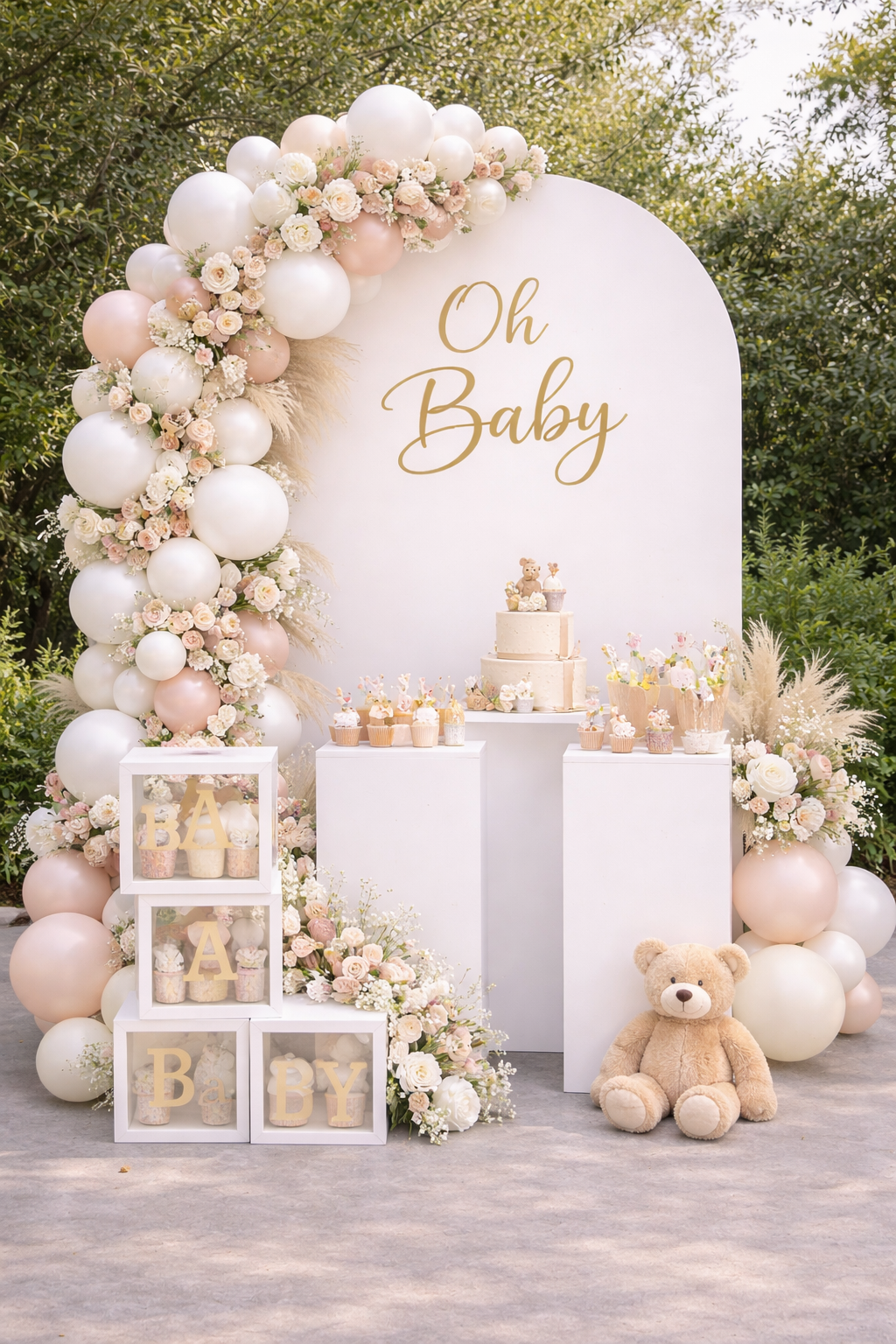 Decorative setup for a baby shower with a large white panel reading "Oh Baby" in gold, surrounded by pink and white balloons and floral arrangements, includes a cake, cupcakes, plush teddy bear, and baby-themed decorations.