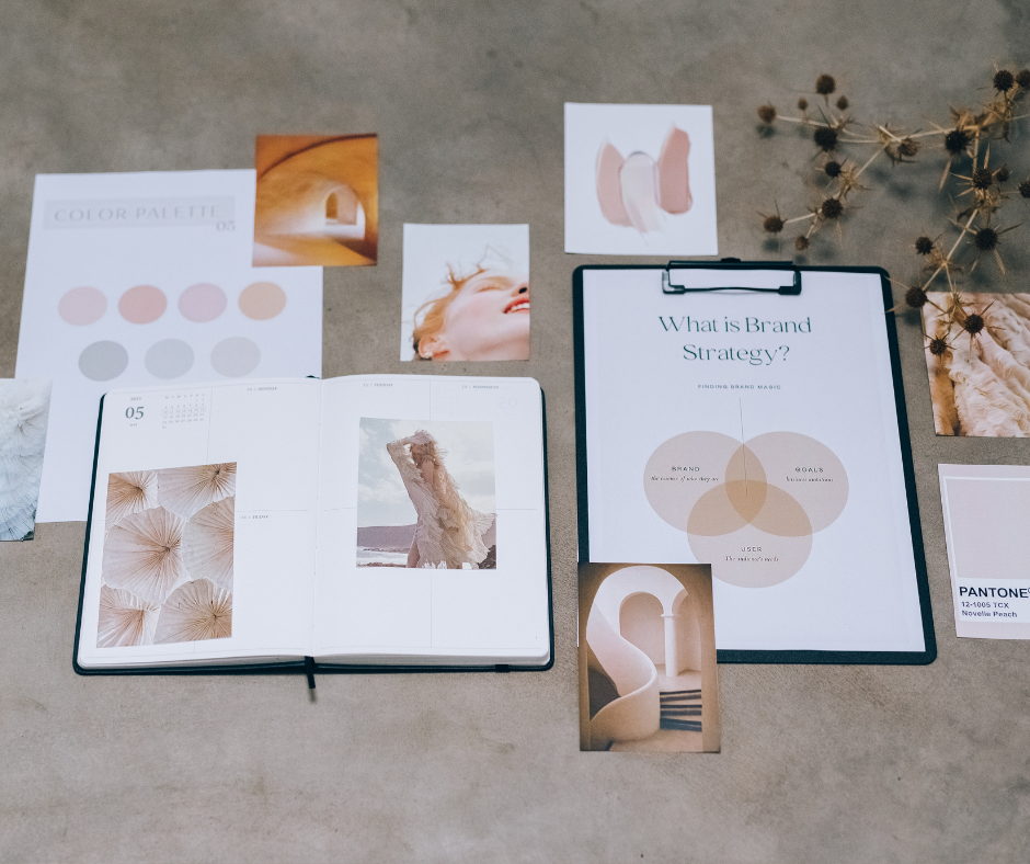 A workspace with open notebooks, color palettes, photographs, art cutouts, and a clipboard with a brand strategy diagram on a beige surface.