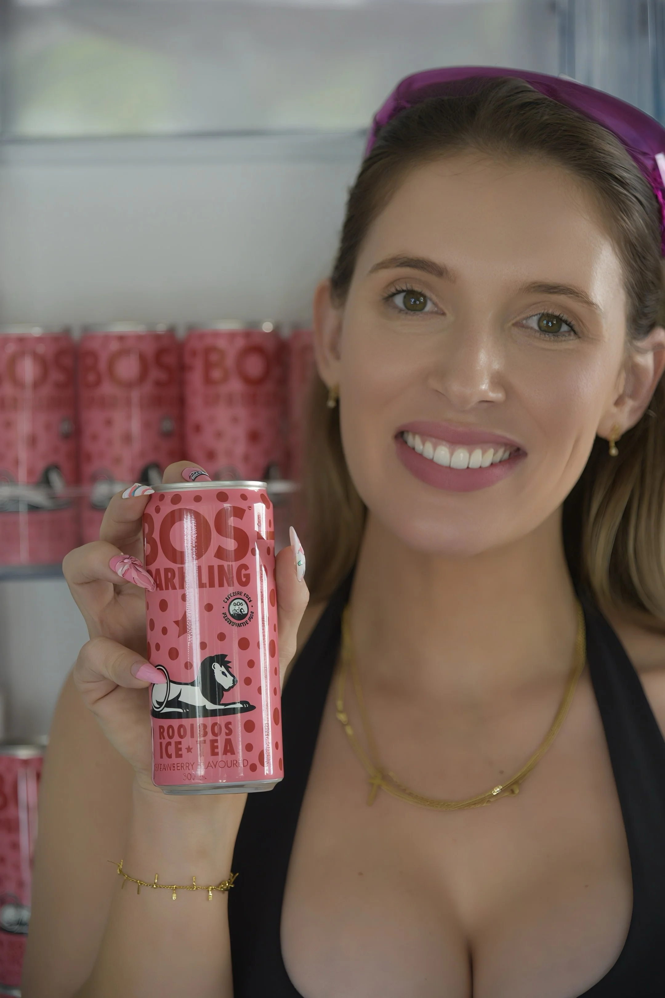 A woman holding a pink can of Rooibos Ice Tea with a lion illustration, smiling, in front of a display of more cans at a store.