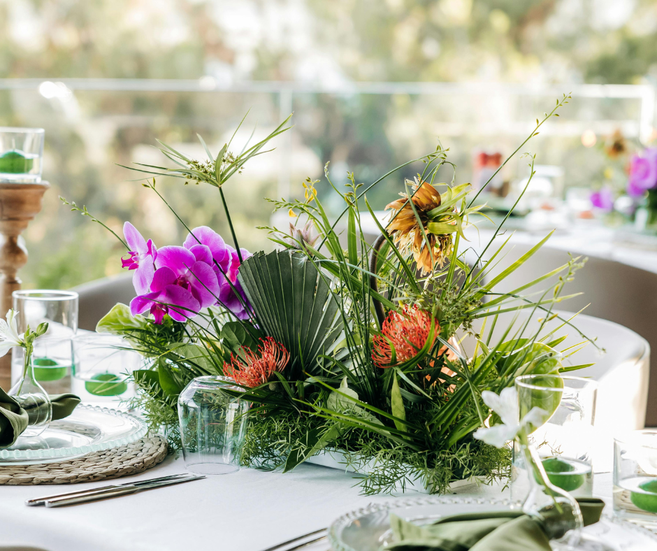 A floral centerpiece with pink orchids, orange protea, sunflower, and assorted greenery on a white table setting.