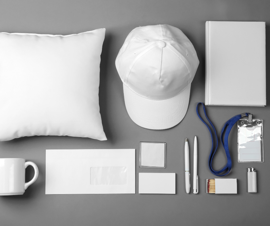 Assorted white office supplies including a pillow, baseball cap, clipboard, notebook, mug, 3 boxes, and pens, on a gray surface with a gray background.