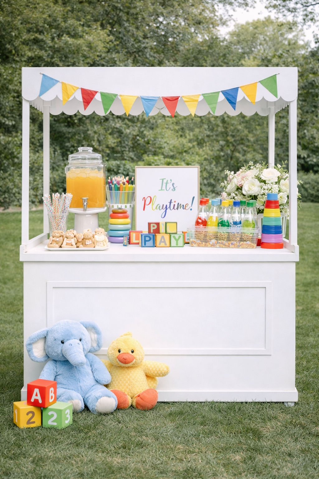 A colorful outdoor party setup with a white cart decorated with a striped banner. The cart has drink dispensers, paper cups, toys, and a framed sign saying 'It's Playtime!'. Stuffed elephants, a duck, building blocks, and toys are arranged on and around the cart, set against a background of green trees and grass.