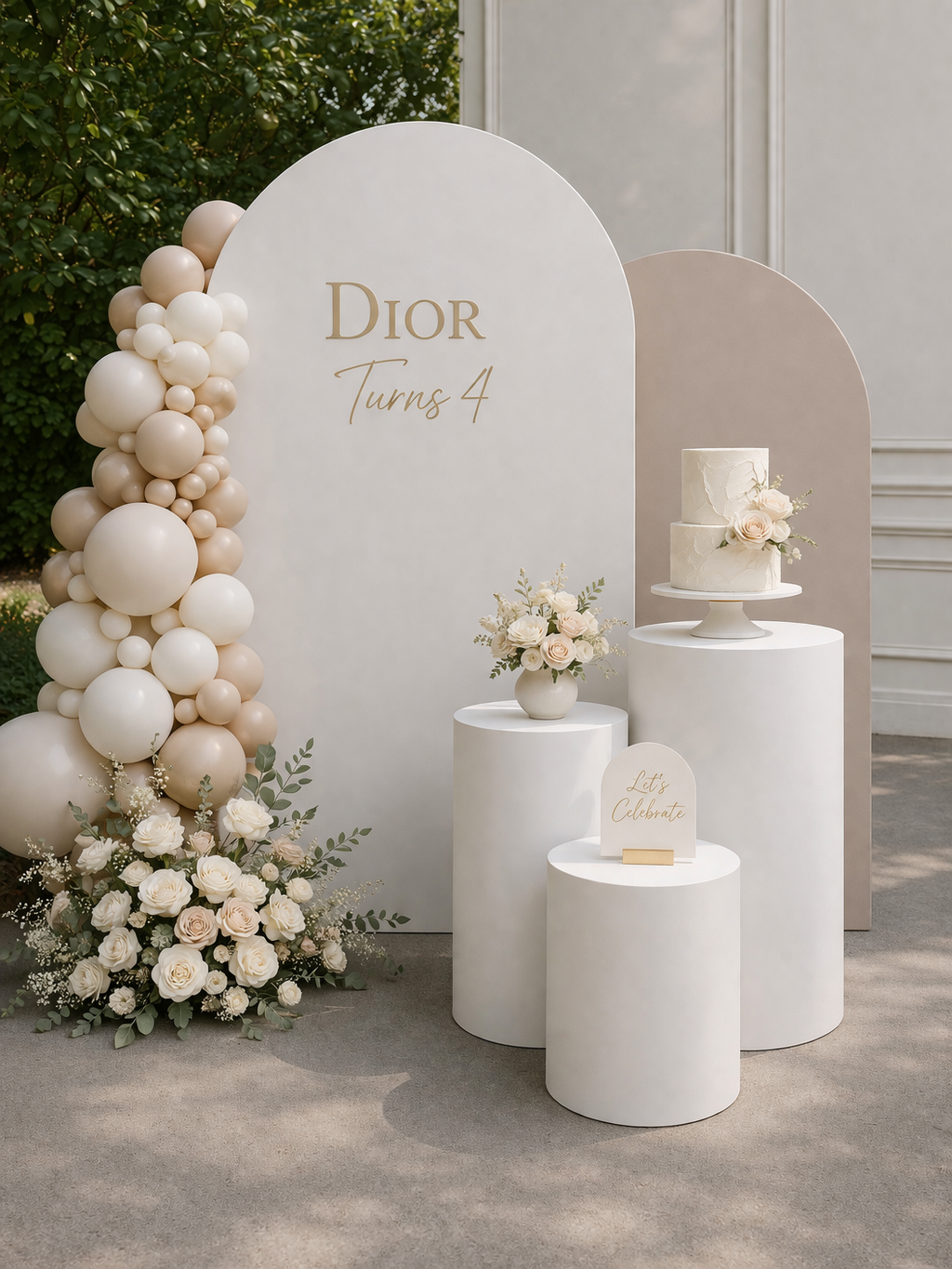 A minimalist celebration setup with a large white arch backdrop that says "Dior Turns 4" in gold lettering, decorated with a balloon arch in shades of white and beige, a bouquet of white roses and greenery, and two white cylindrical pedestals, one with a white floral cake and the other with a small sign that says "Let's Celebrate".
