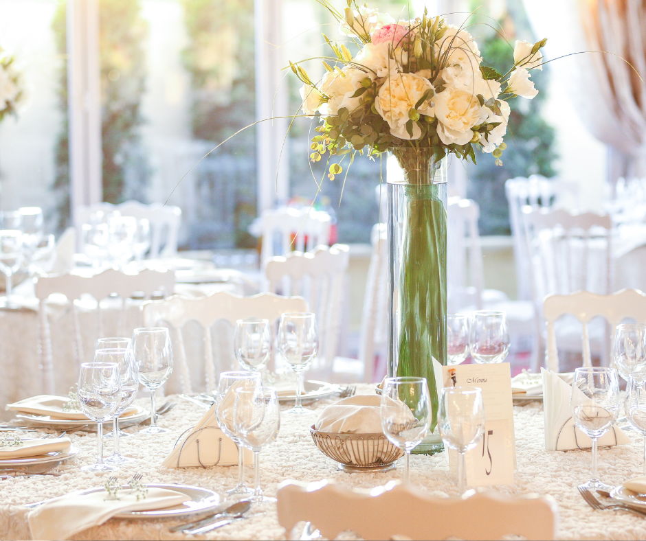 Elegant banquet table decorated with a tall floral centerpiece of white and cream-colored roses and greenery in a tall glass vase. The table is set with wine glasses, plates, napkins, and a small menu card. The setting appears for a formal event in a