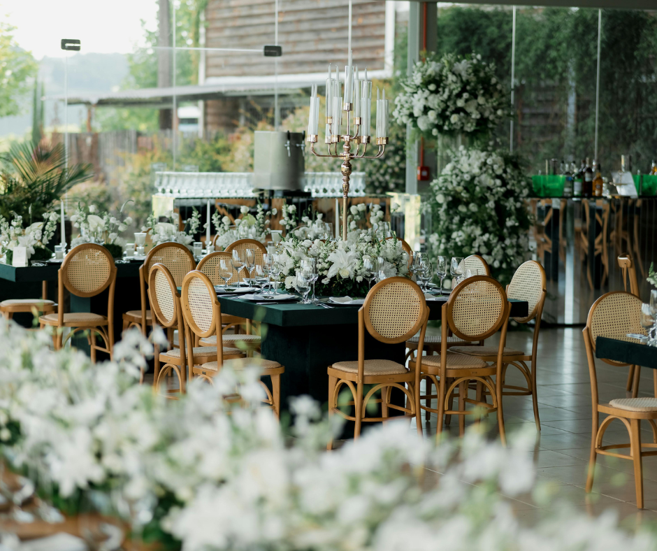 Elegant banquet table set with white floral centerpieces, surrounded by wooden chairs with woven rattan backs, featuring tall candelabra centerpiece in a well-lit venue with large windows and greenery outside.