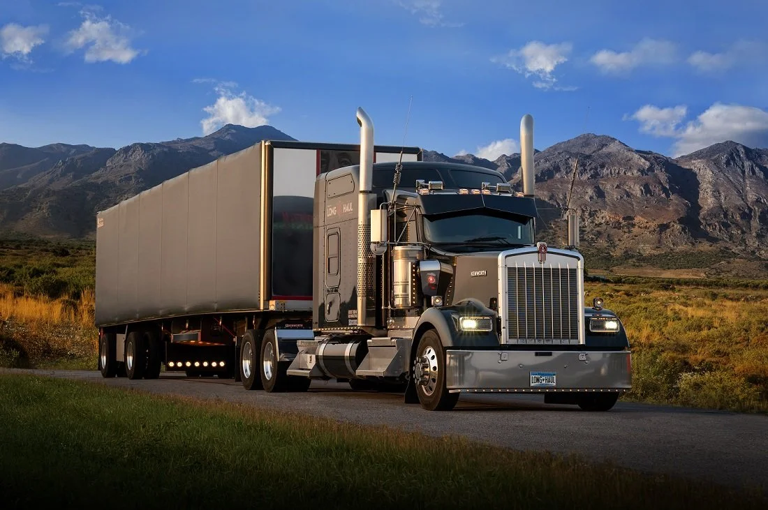 A large black semi-truck with a silver trailer driving on a rural road with mountains in the background under a partly cloudy sky.