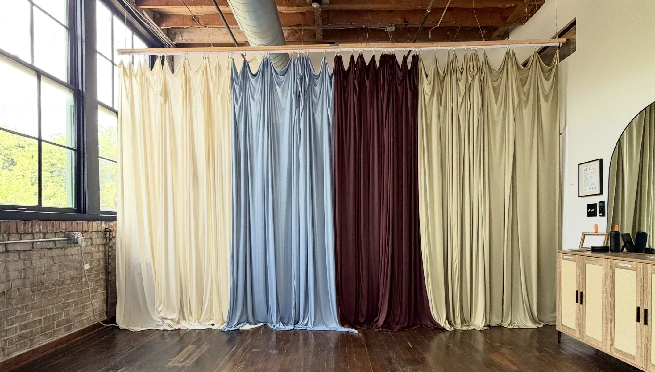 Colorful curtains hanging from a ceiling track in a room with brick wall and large window.