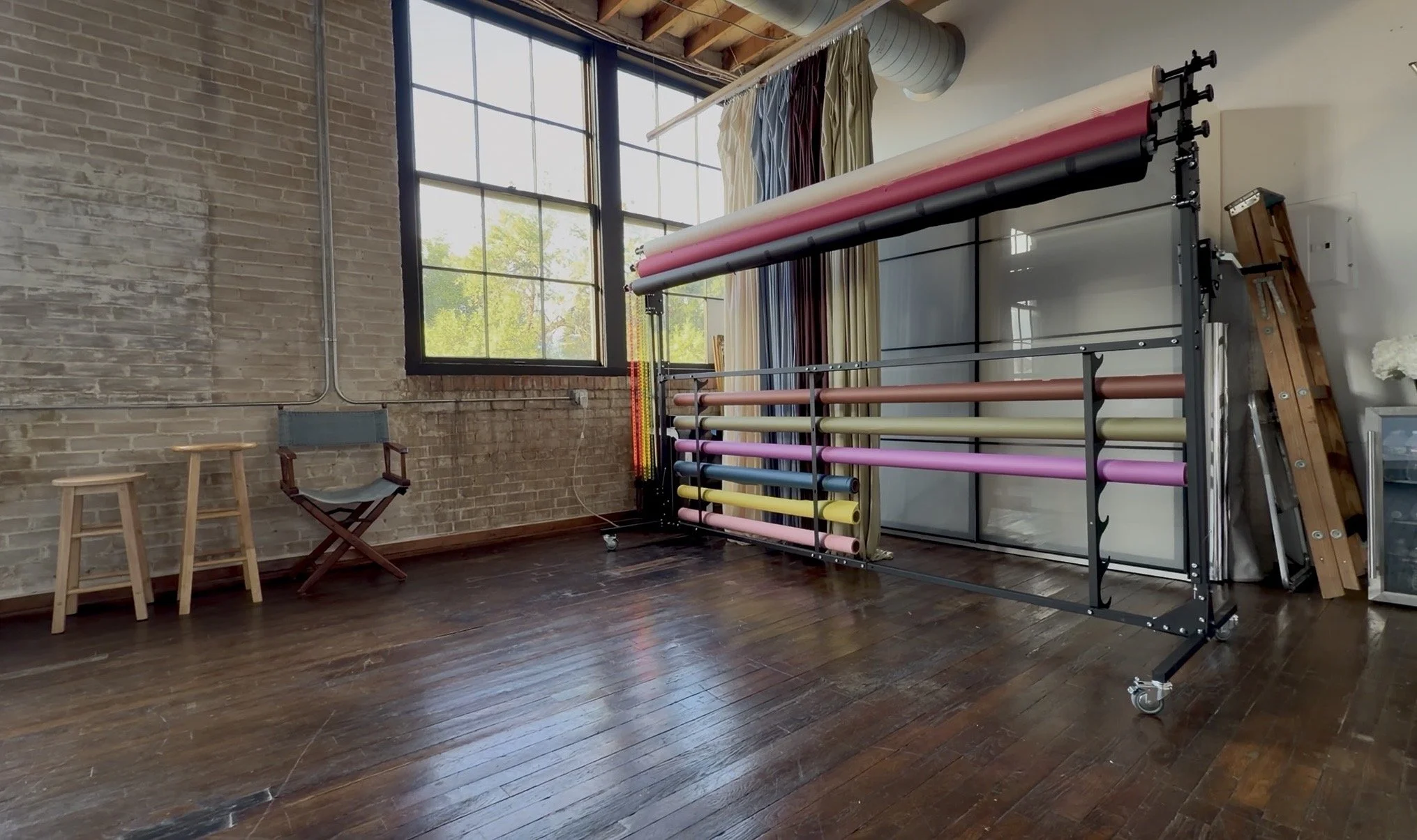 A room with exposed brick walls and large window, featuring a clothing rack with multiple rolls of colorful fabric, wooden stools, and a director's chair.