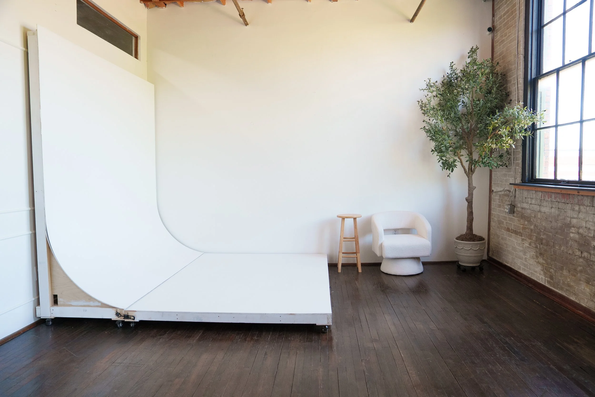An empty photography studio with a white backdrop, a small wooden stool, a white chair, and a tall potted plant near a large window with black frames and an exposed brick wall.
