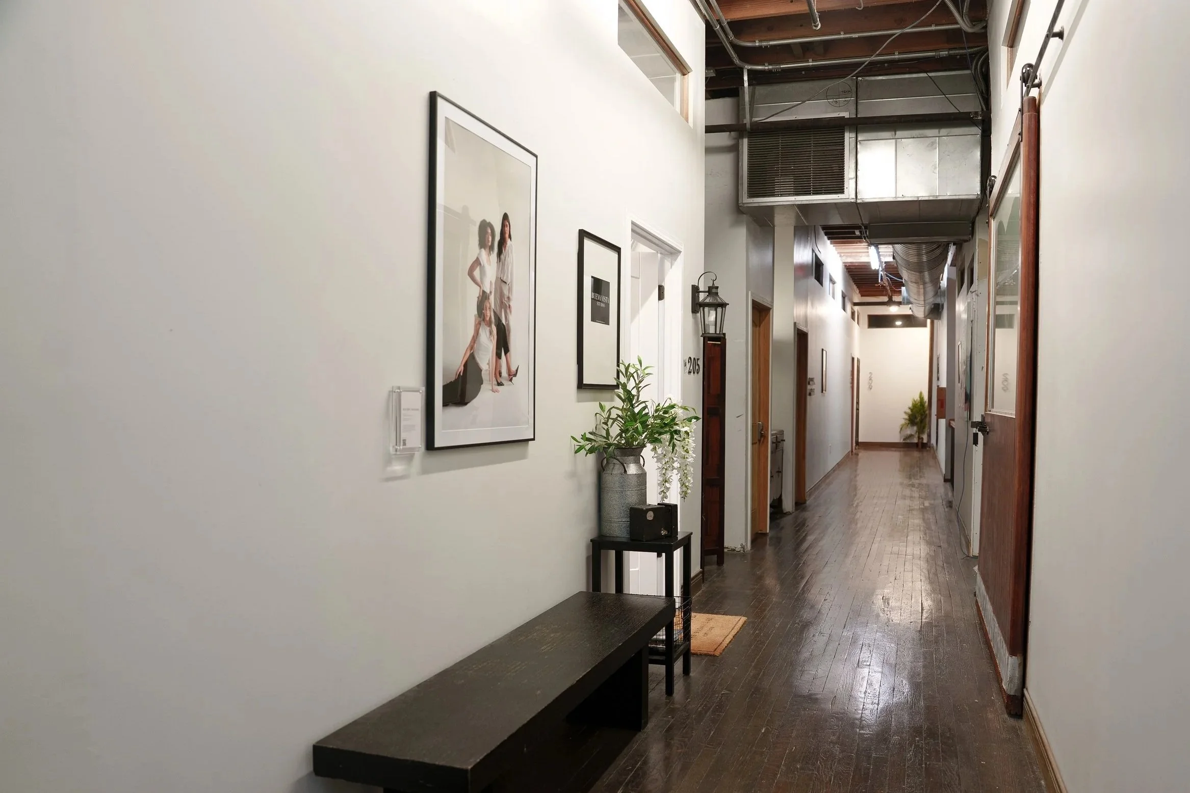 A hallway with wooden flooring and white walls, decorated with framed photographs, plants, and wall-mounted lights. Overhead pipes and ductwork are visible on the ceiling.