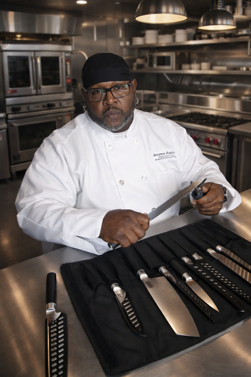 Chef with black head covering and glasses sitting at a stainless steel table with a set of knives in a black case, holding a large knife in a professional kitchen.