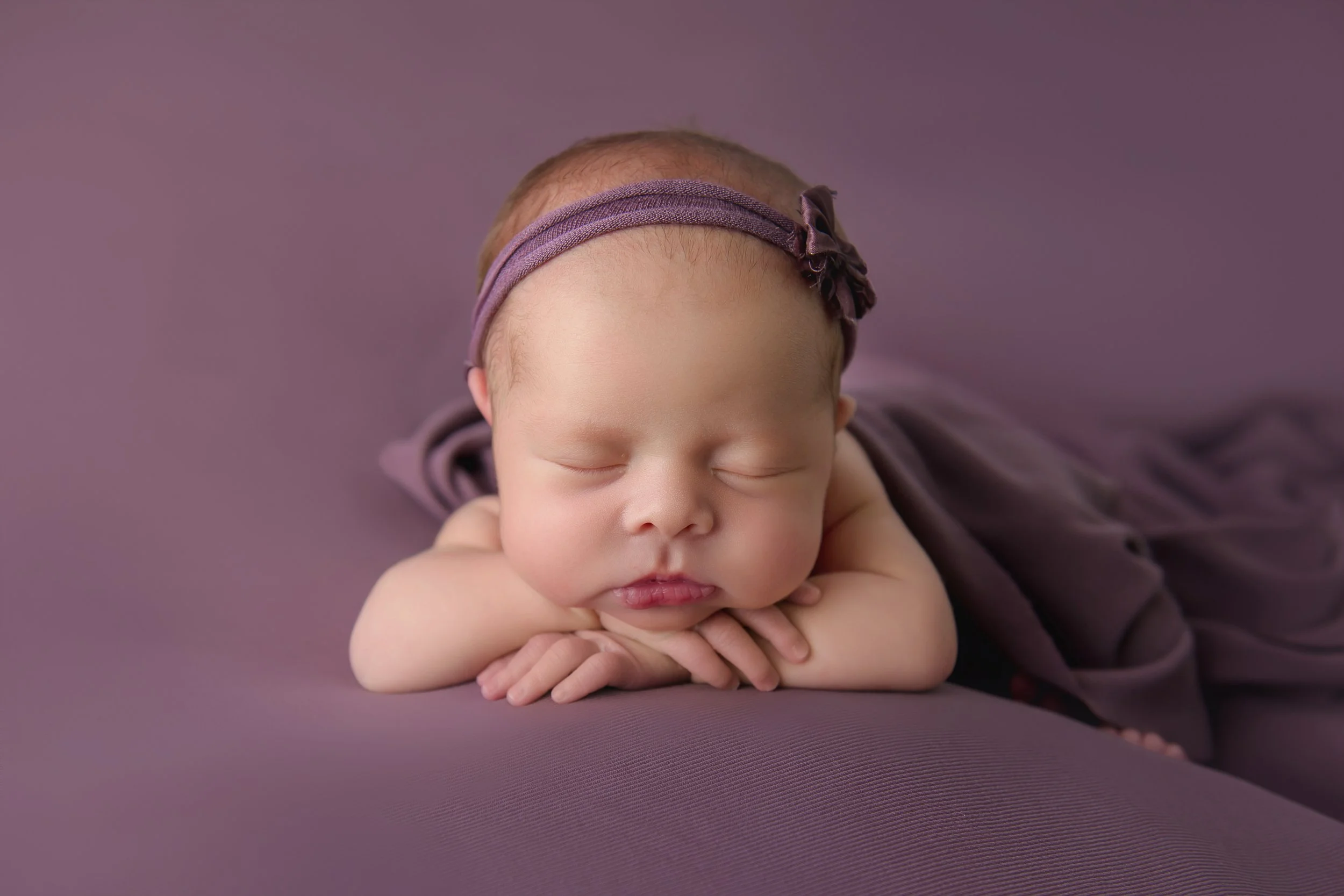Fine art newborn portrait of a sleeping baby with a purple headband, styled on layered purple textures in our luxury studio.