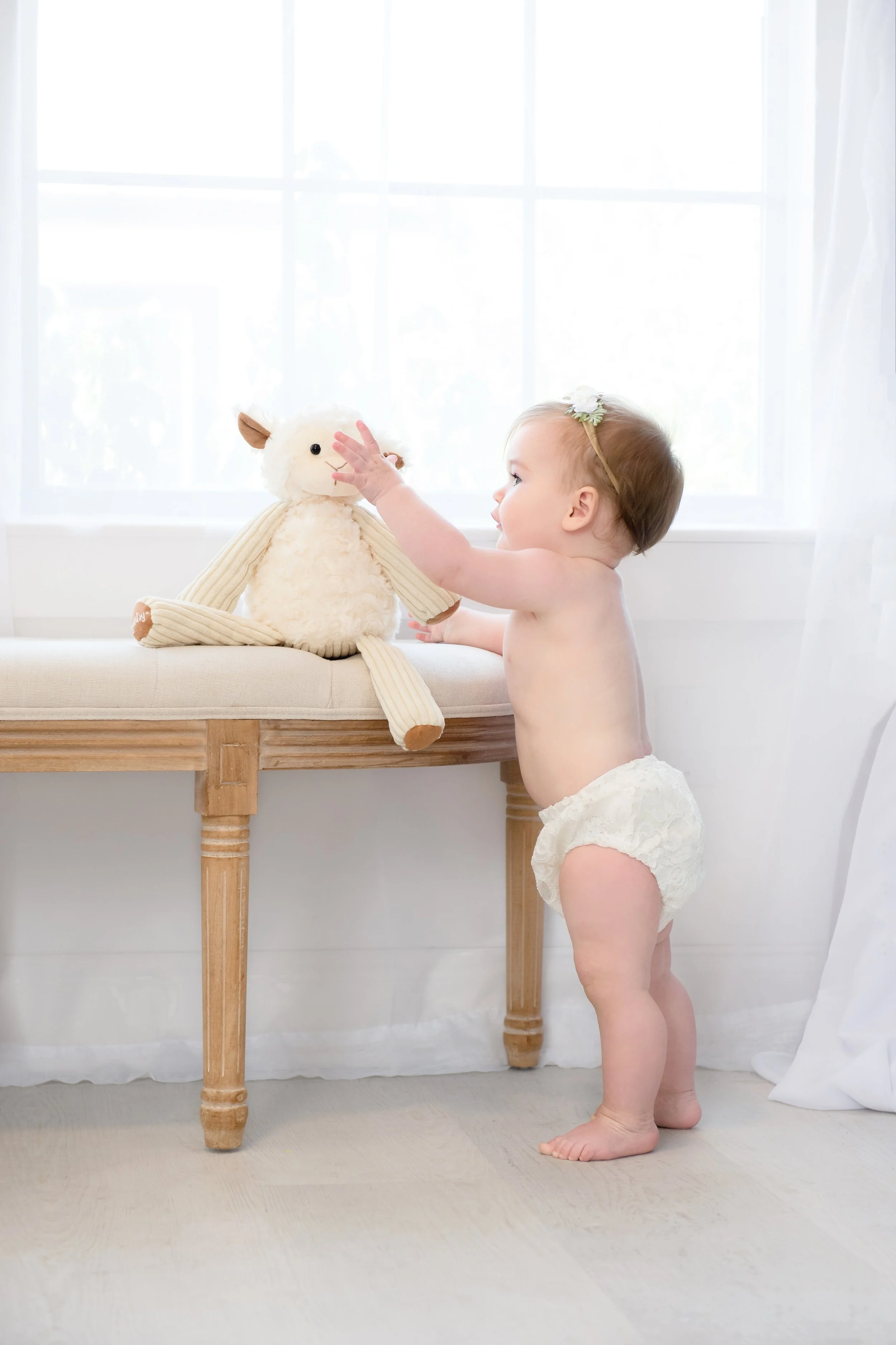 A baby in a diaper playing with a plush stuffed animal on a wooden bench near a window with sheer white curtains.