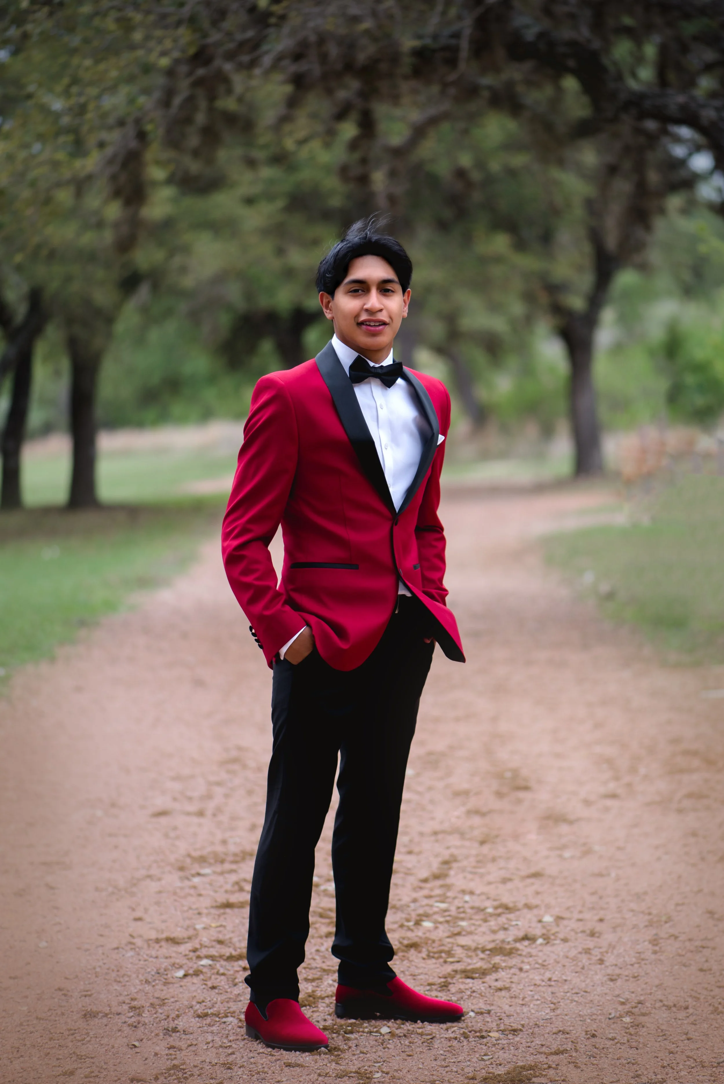 A young man in a red tuxedo with black lapels, wearing a white shirt, black bow tie, and red shoes, stands on a dirt path in a park with trees in the background.