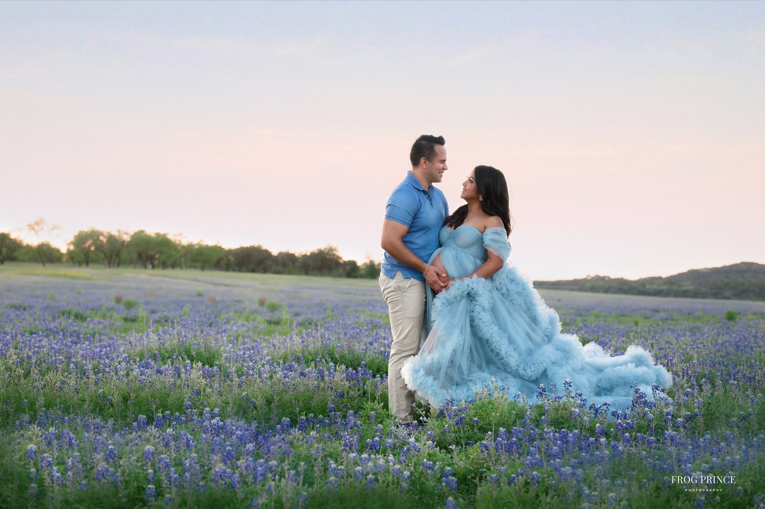 Luxury maternity portrait of a couple standing in a field of purple flowers at sunset, with the mother in a flowing blue gown and her partner holding her baby bump in the Hill Country near Spring Branch.