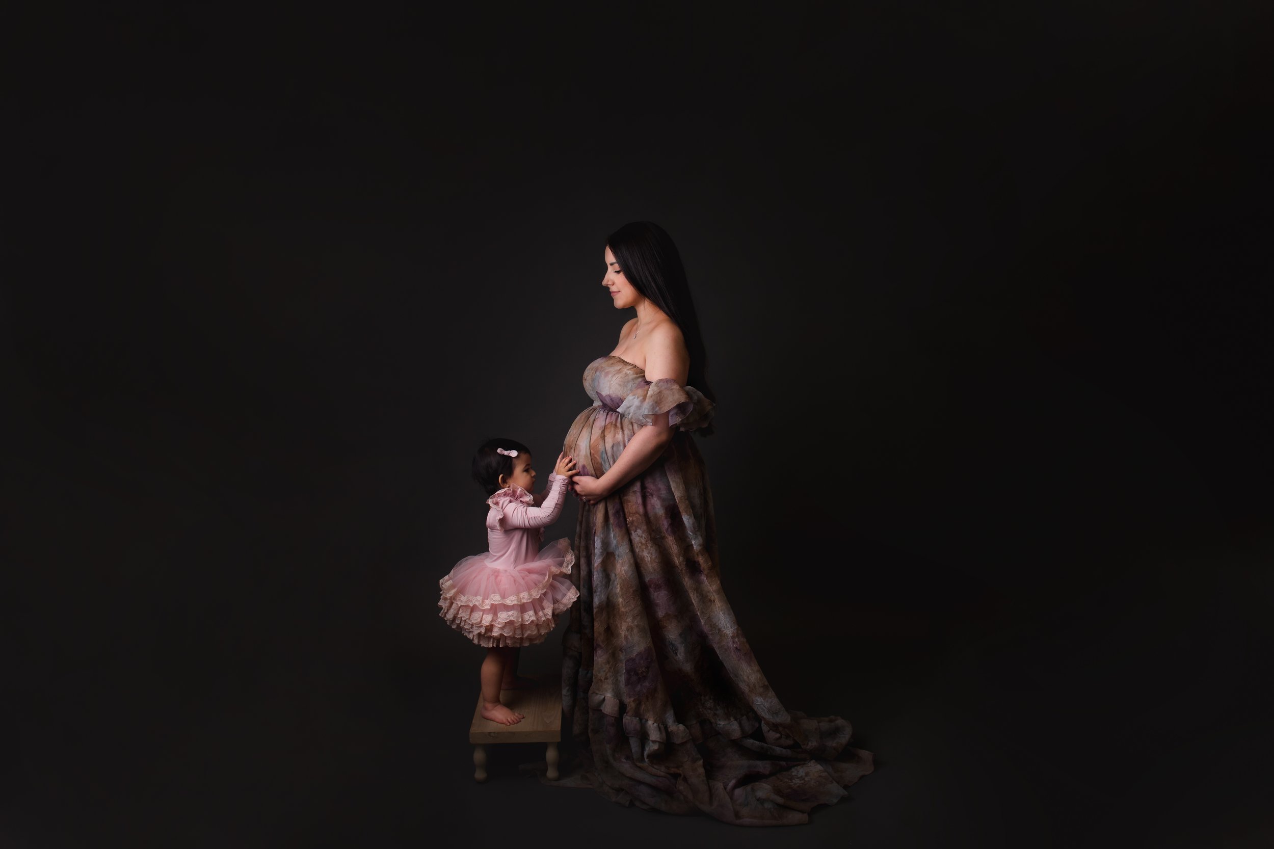 Luxury maternity portrait of a pregnant mother in an elegant patterned gown standing on a studio platform, with her young daughter in a pink tutu gently touching her baby bump against a dramatic dark background.