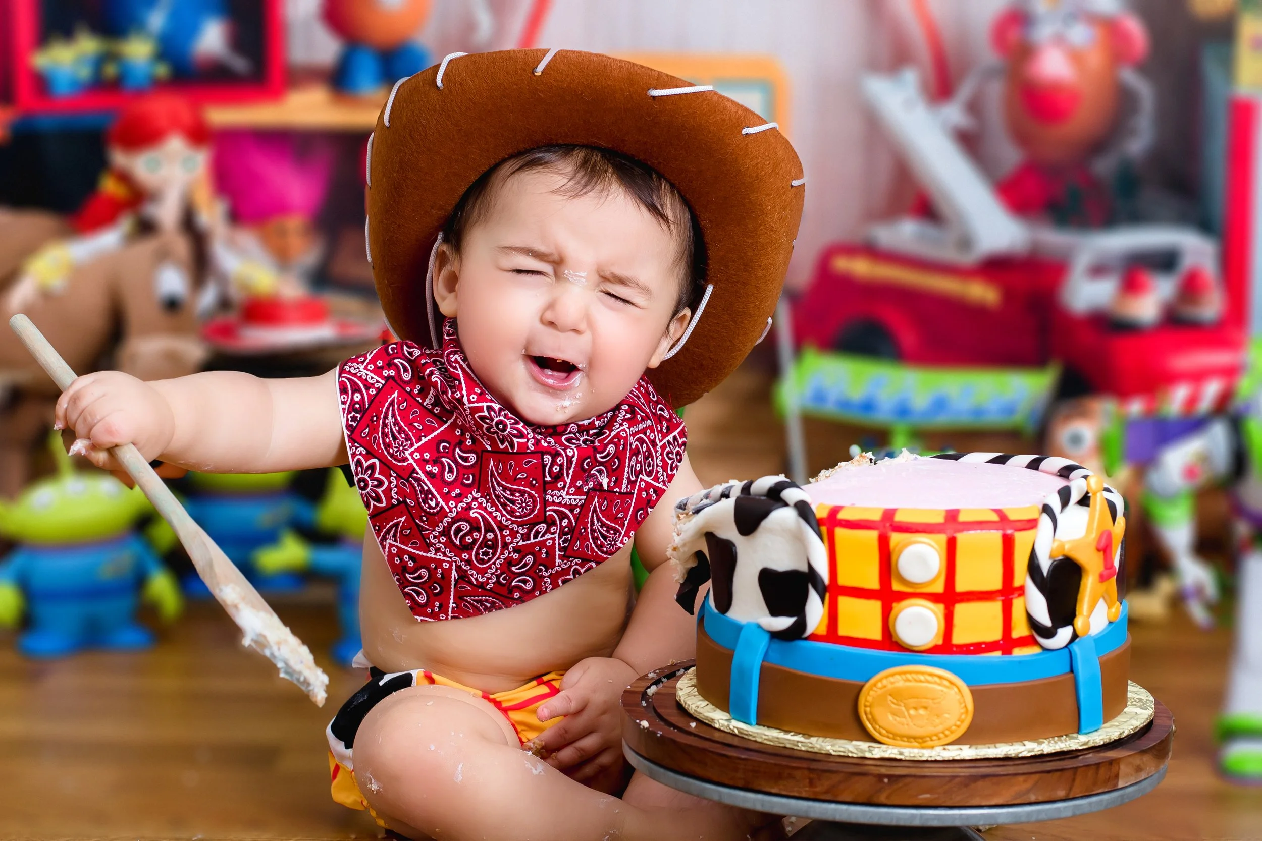 A young child dressed as a cowboy with a brown hat, red bandana, and yellow shorts, sitting at a birthday party with a themed cake in front. The child appears to be upset or crying, with cake and frosting on their face.