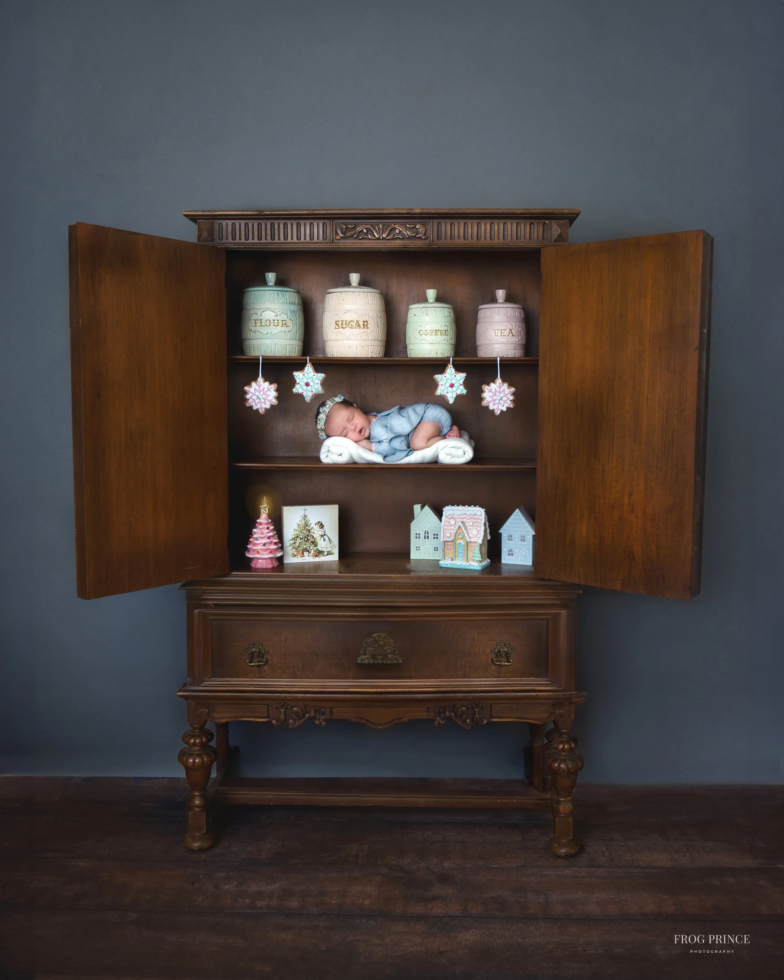 Fine art newborn portrait of a baby in blue resting inside a styled wooden cabinet with vintage baking jars and soft holiday accents in our Spring Branch studio.