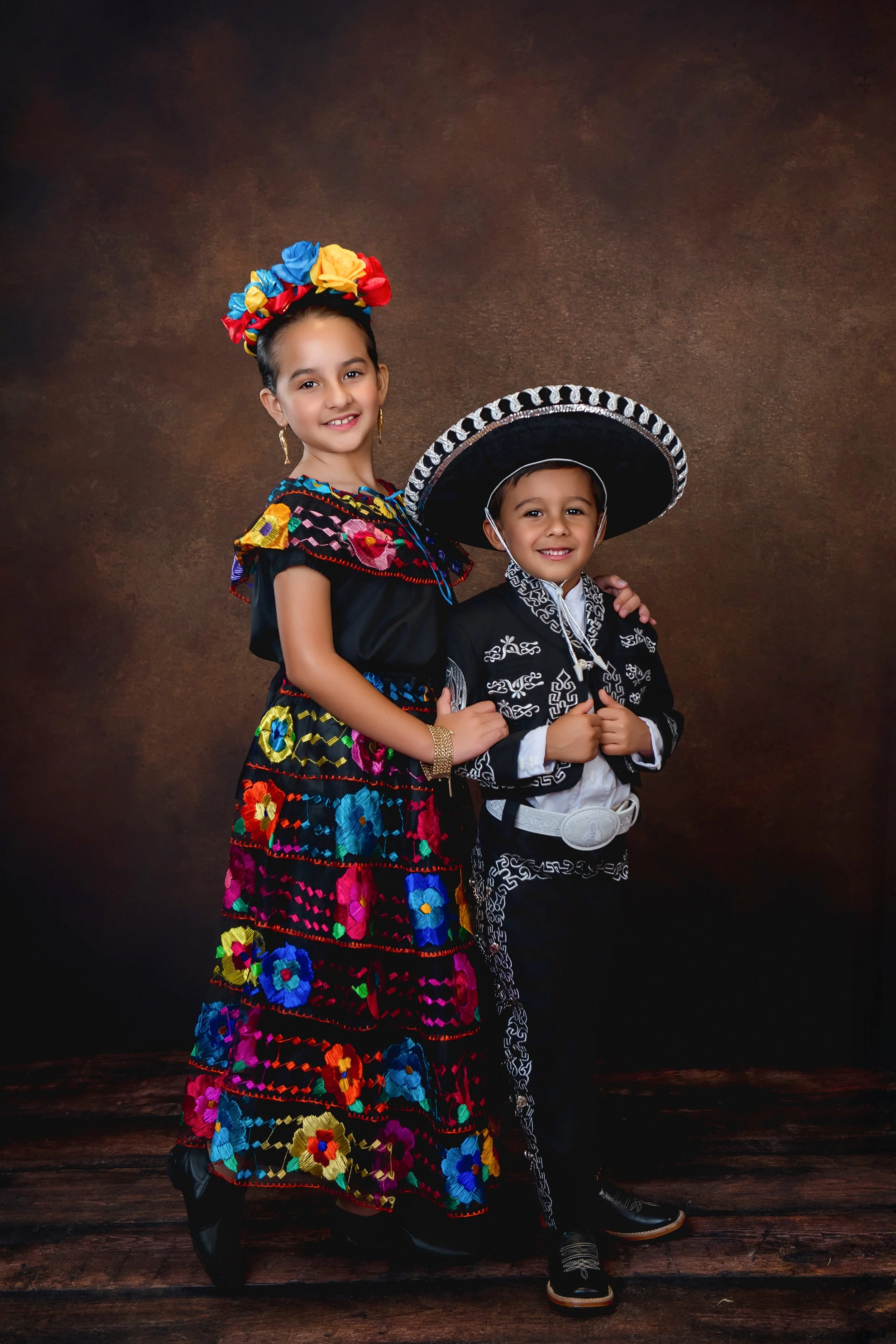 A young girl and boy in traditional Mexican attire, standing together against a brown background. The girl is wearing a colorful embroidered dress with a floral headpiece, and the boy is dressed in a black charro suit with a large sombrero.