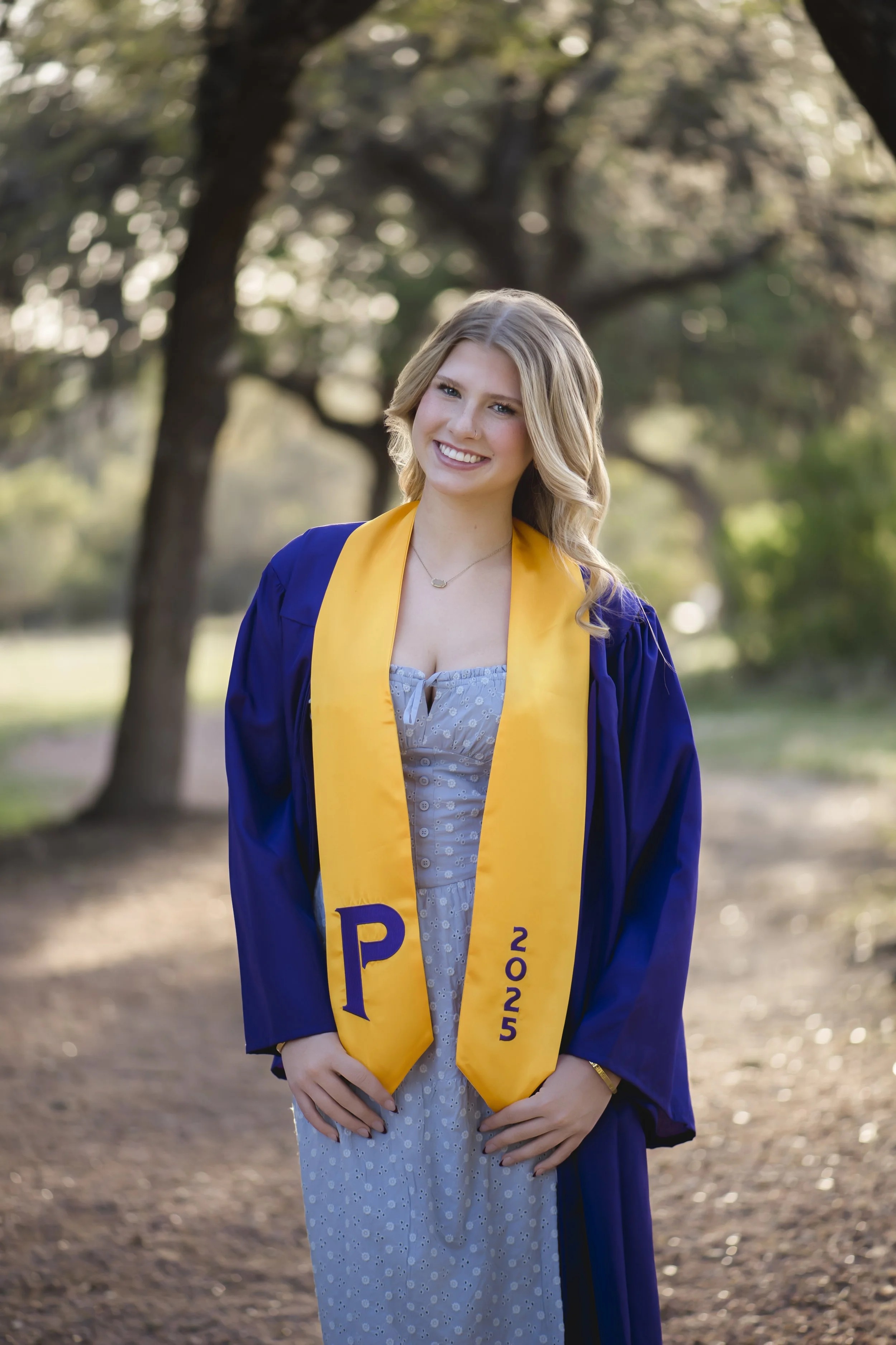 A young woman in a gray dress and blue graduation gown with a yellow stole, standing outdoors on a dirt path surrounded by trees, smiling at the camera.