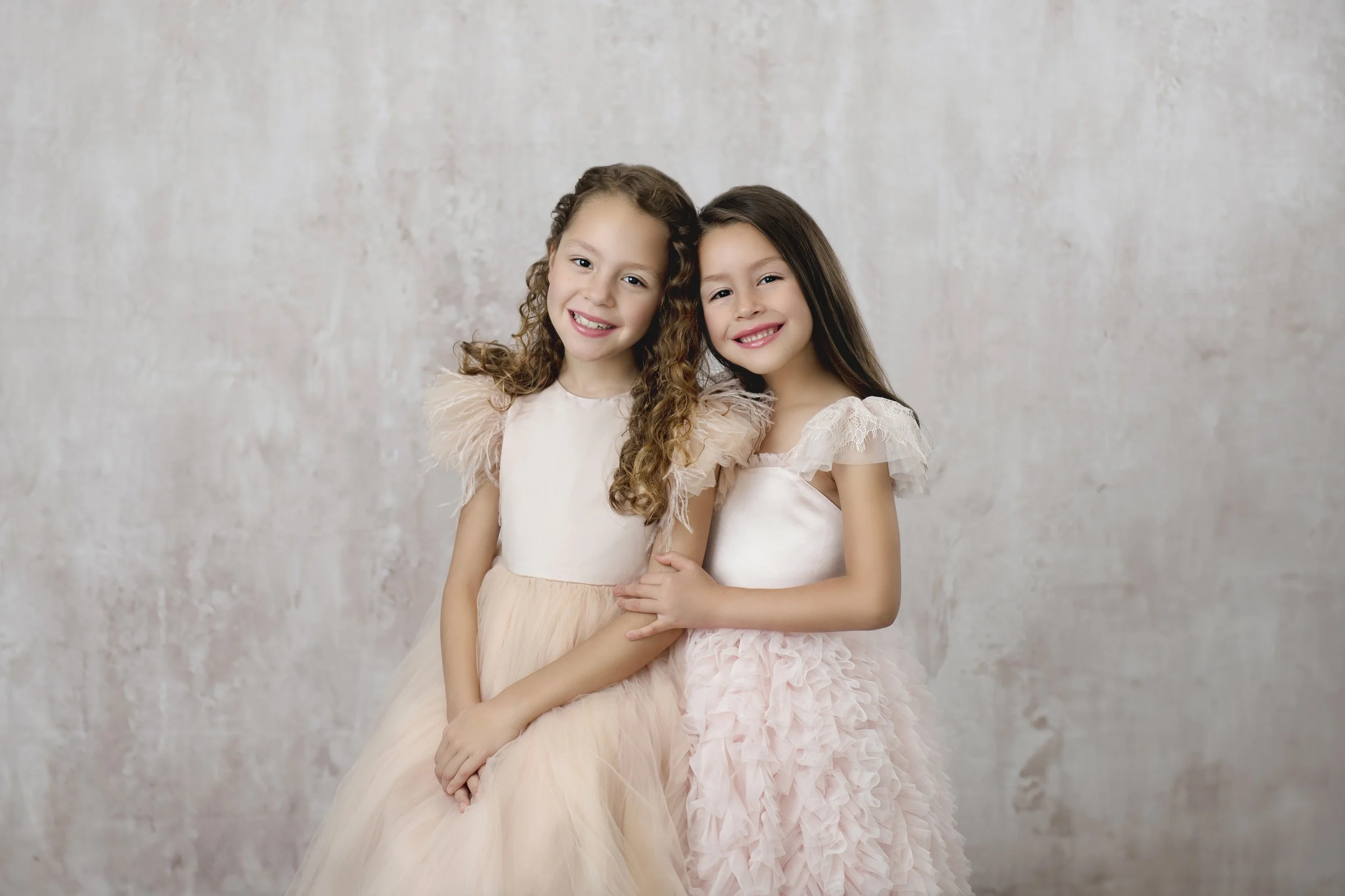 Two young girls in pink dresses smiling and hugging each other in front of a textured light-colored background.