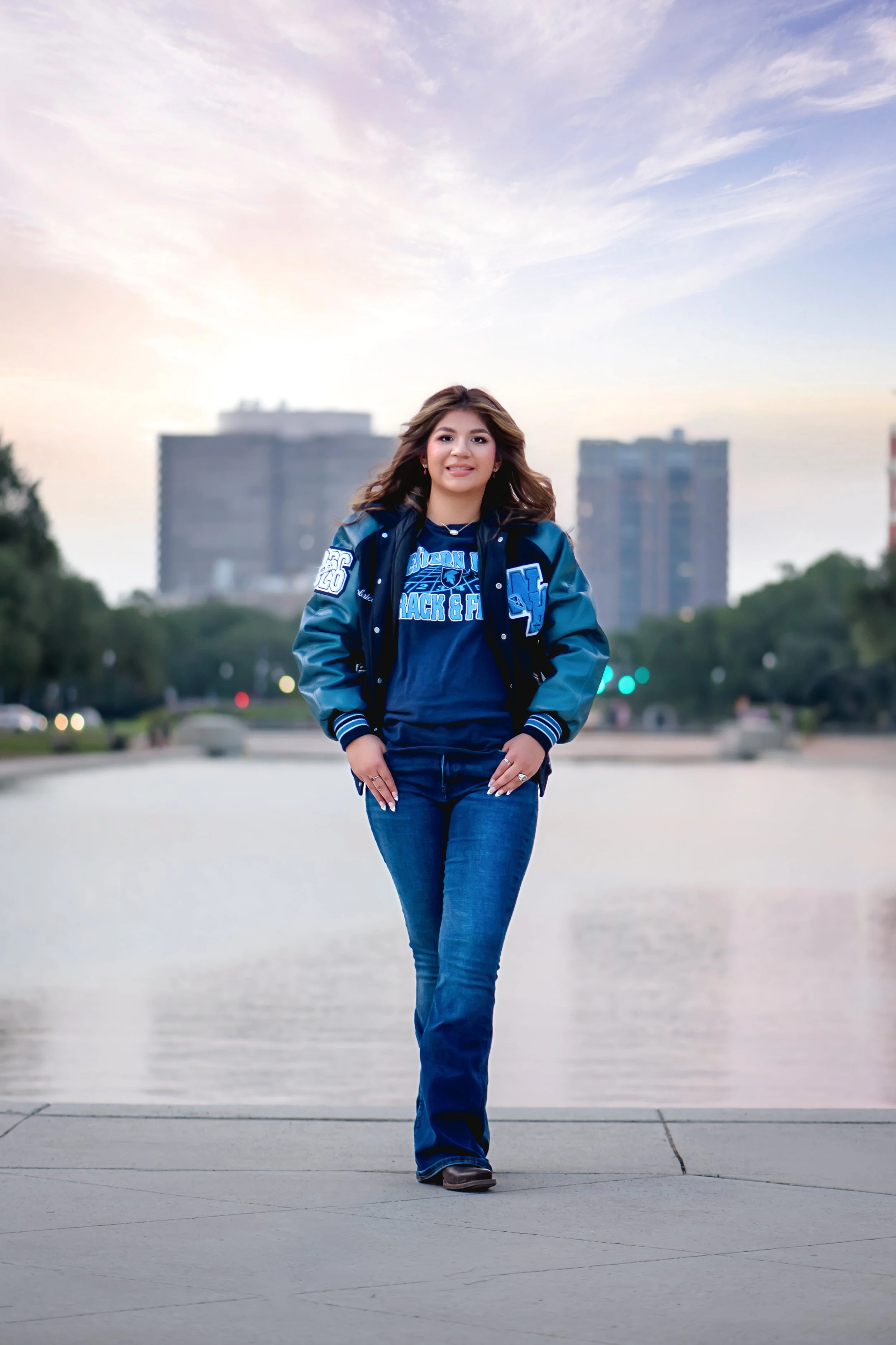 Young woman walking outdoors near water with city buildings in the background during sunset.