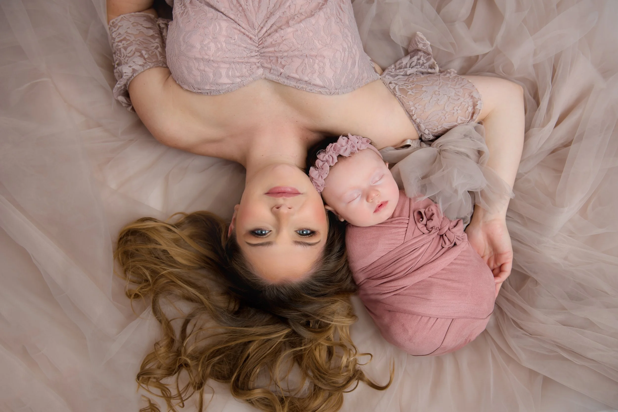 A woman lying on a bed with a baby, both dressed in pink, surrounded by soft pink fabrics. The woman has long, wavy hair and is looking at the camera, while the baby is sleeping with a matching pink headwrap in our Spring Branch studio.