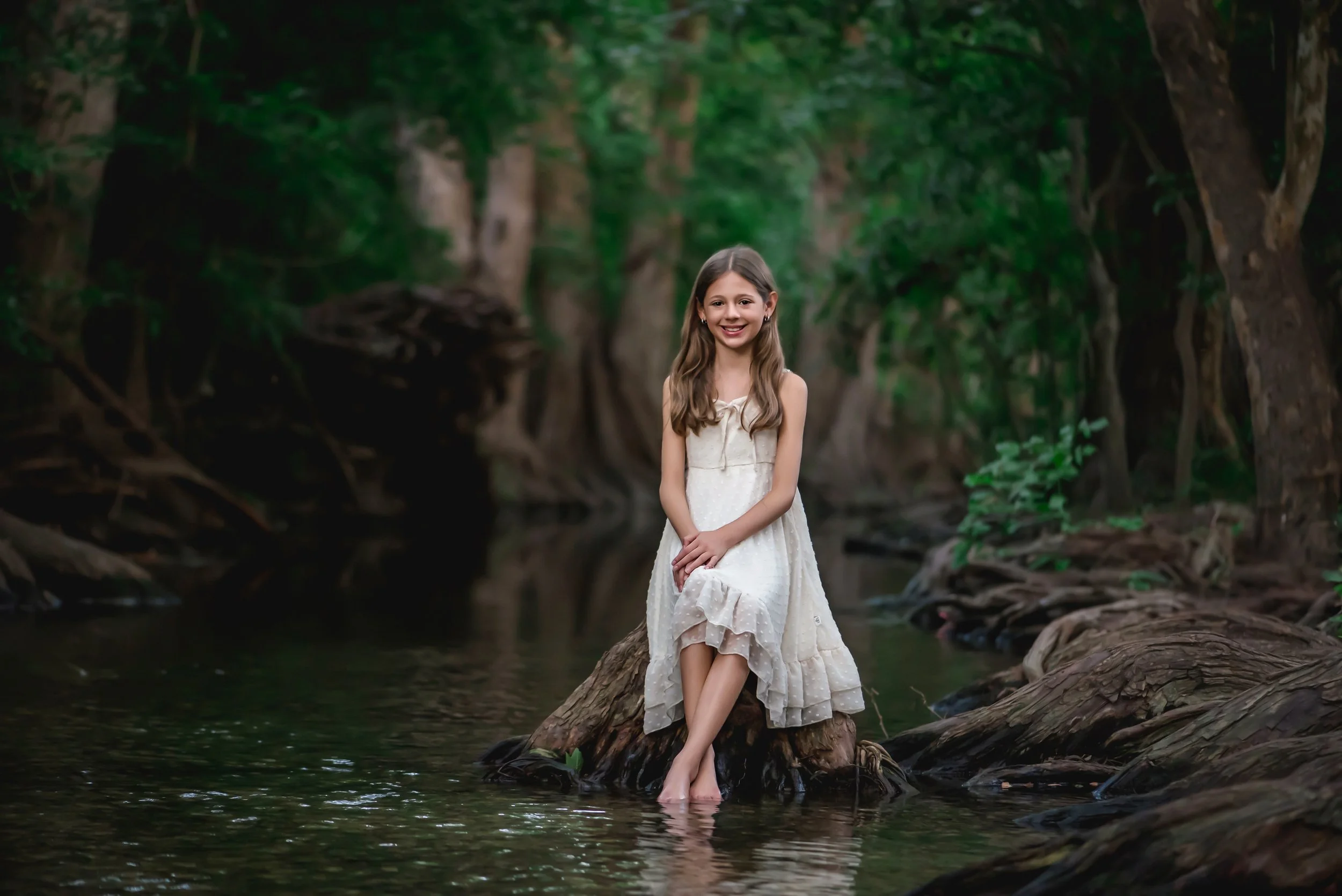 A young girl in a white dress sitting on a fallen log in a forest stream, smiling at the camera.