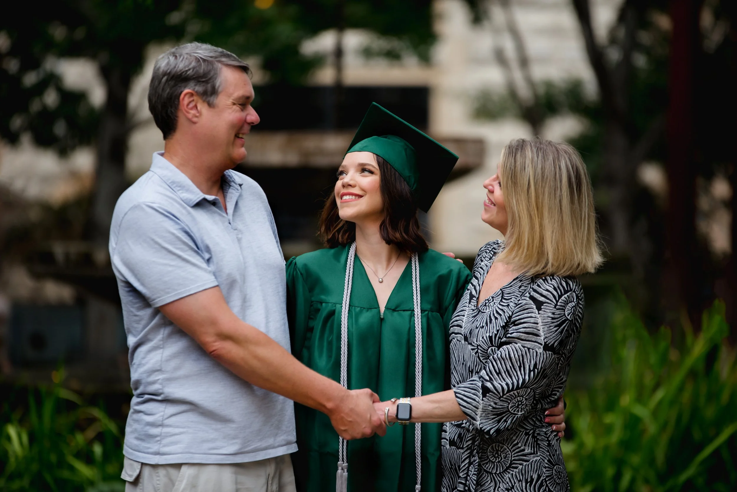 A young woman in a green graduation gown and cap is smiling and shaking hands with a man, while a woman in a patterned dress looks on and smiles in an outdoor setting with greenery.