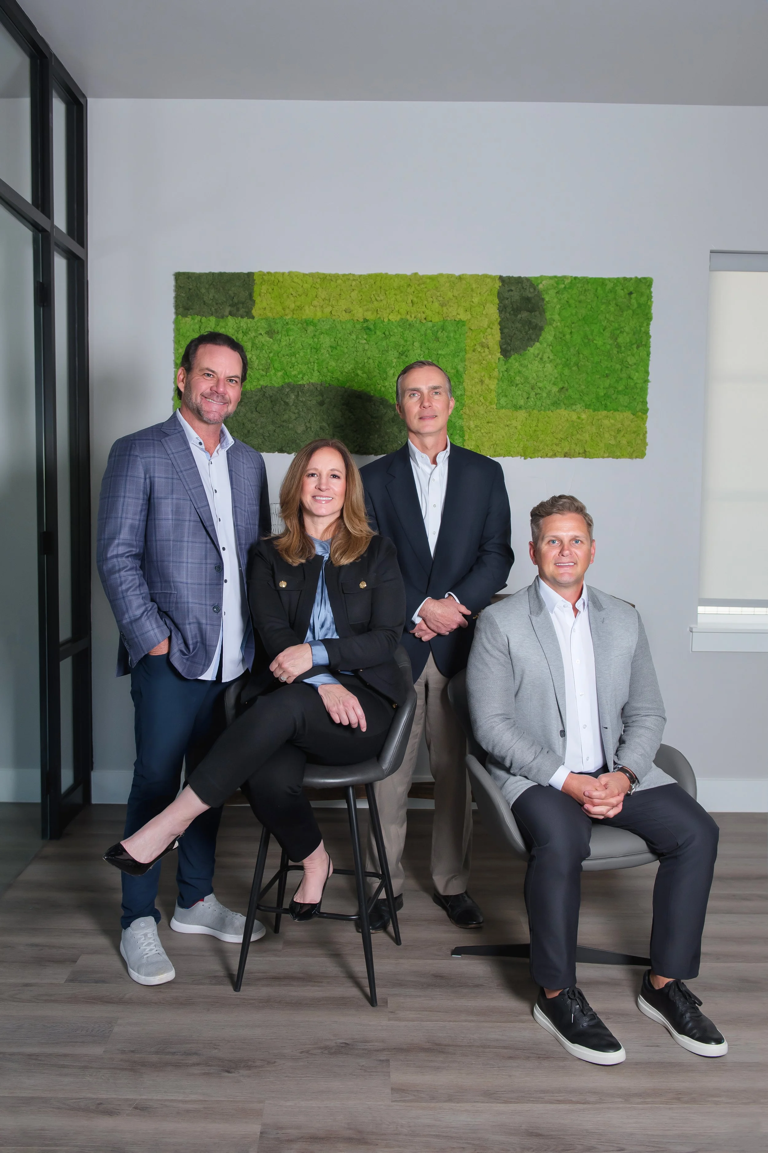 Group of four business professionals in an office, two men standing and two seated women, with a green abstract wall art in the background.