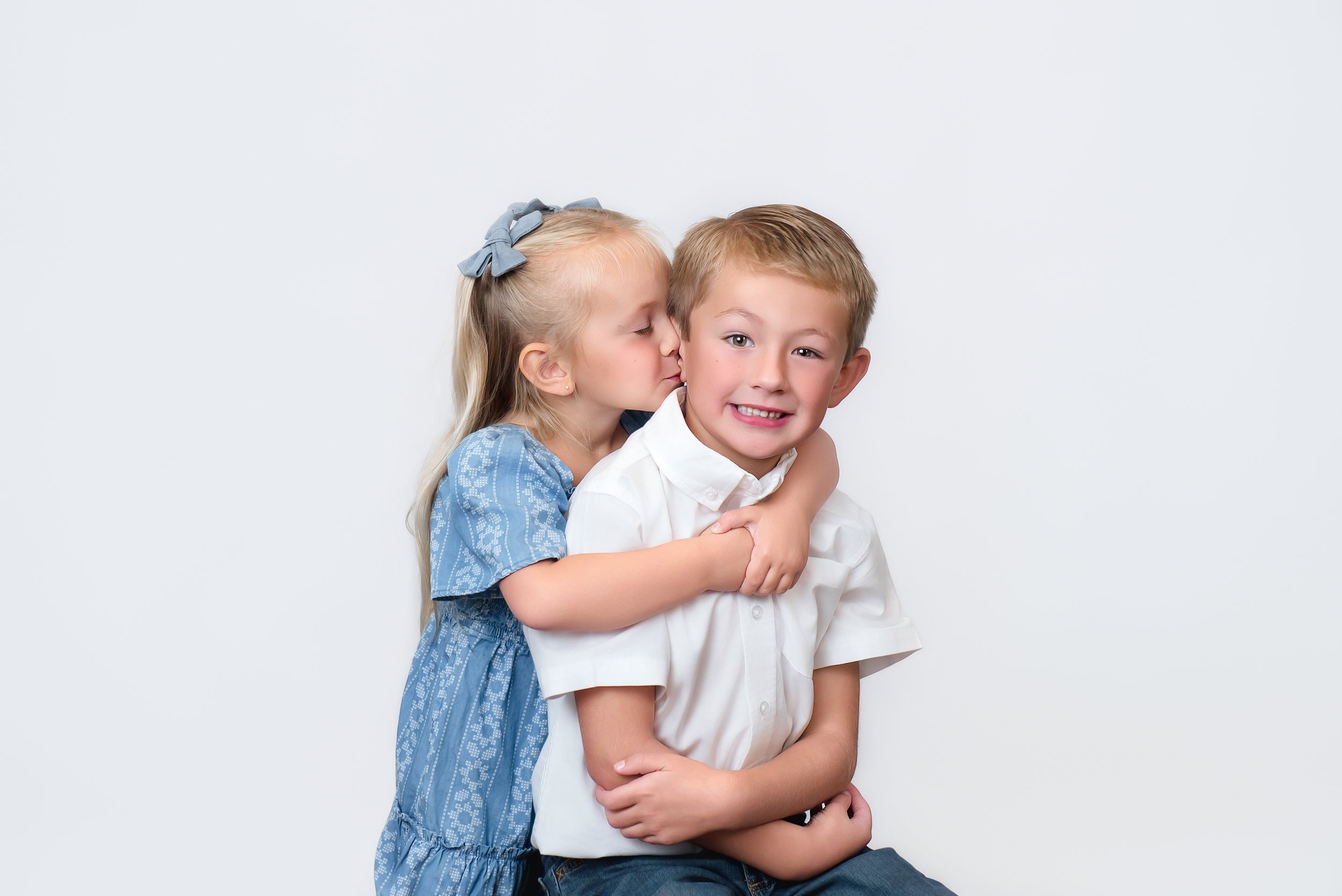 A young girl with blonde hair and a blue dress leaning in to kiss a smiling young boy with a white shirt on a white background.