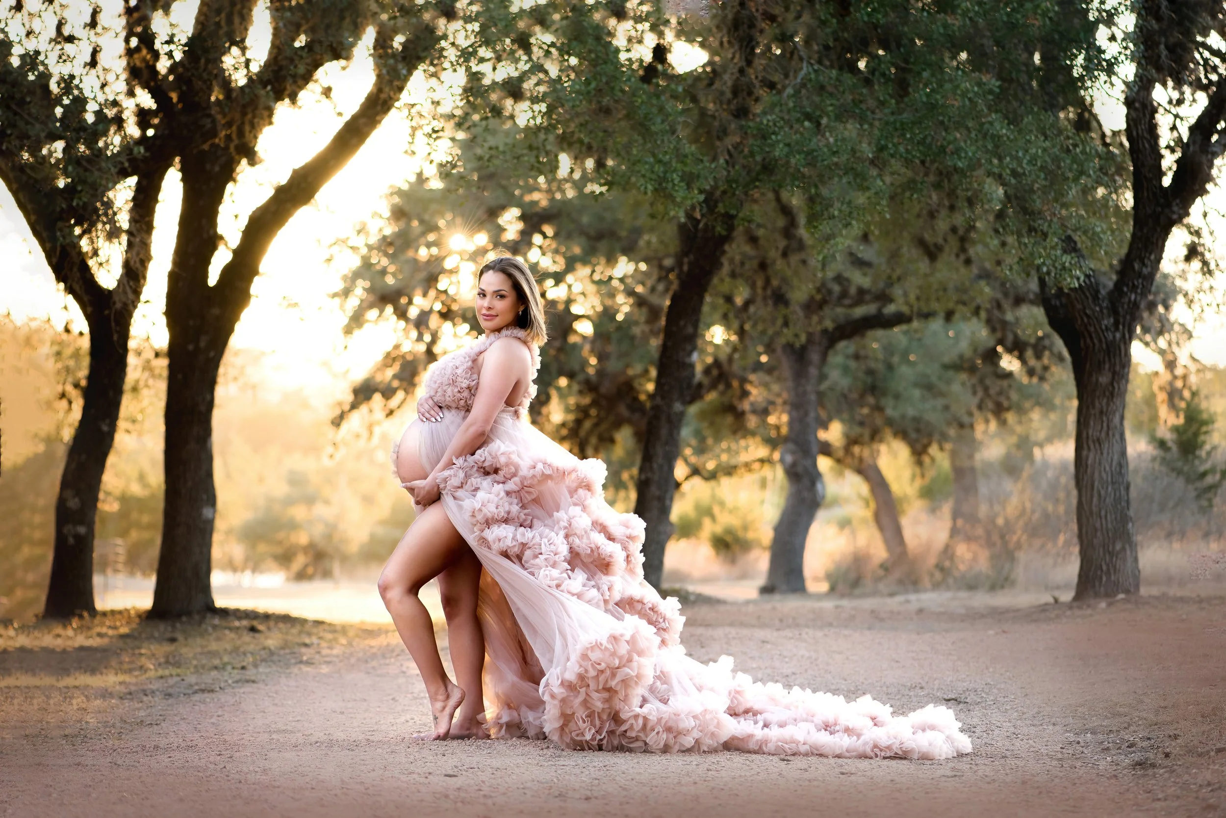 Fine art maternity portrait of a pregnant woman in a flowing pink gown standing on a dirt path at sunset in a Hill Country park near Bulverde.
