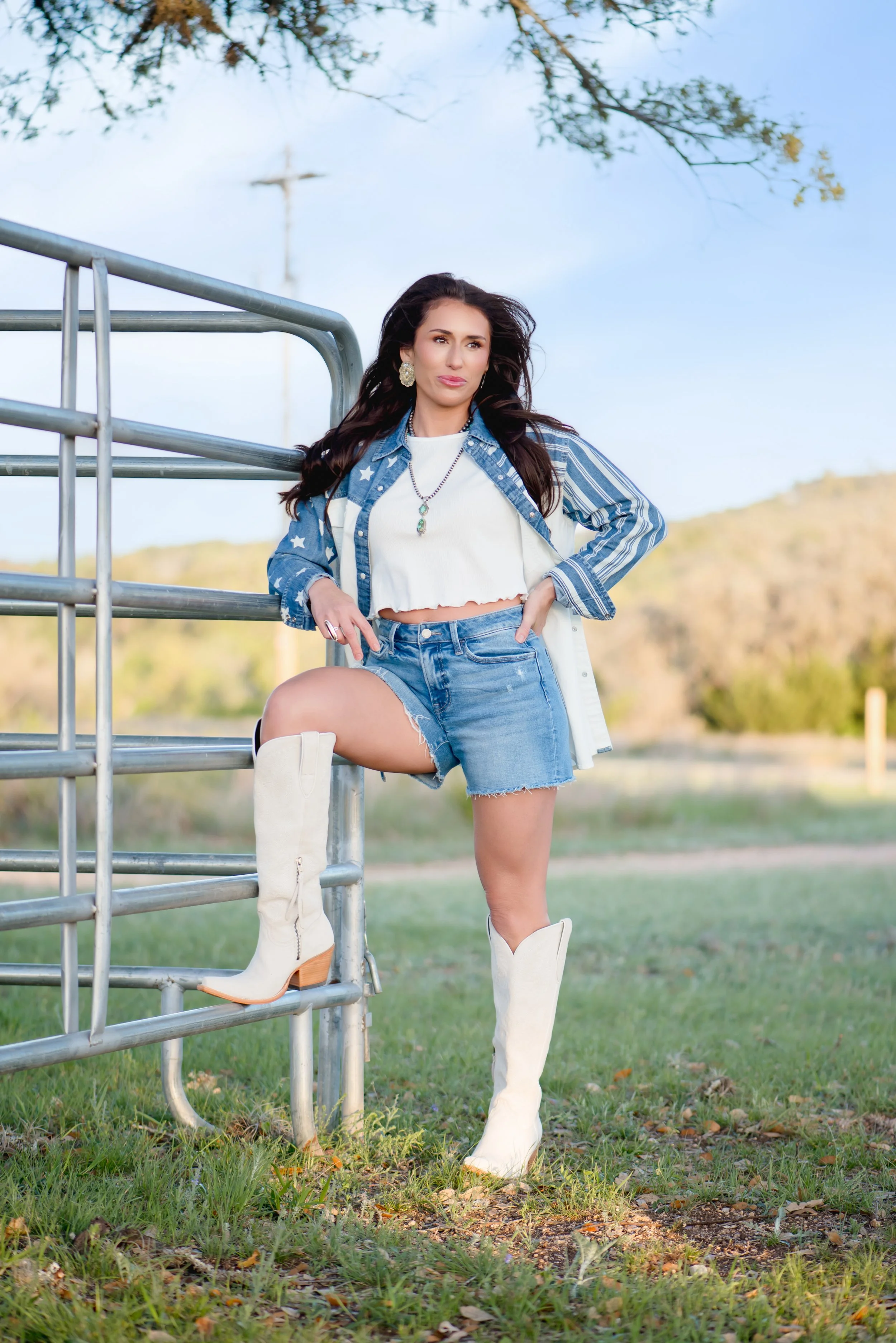A woman standing outdoors with one foot on a metal fence, wearing denim shorts, a white crop top, a denim jacket, and white knee-high cowboy boots, with a scenic landscape in the background.