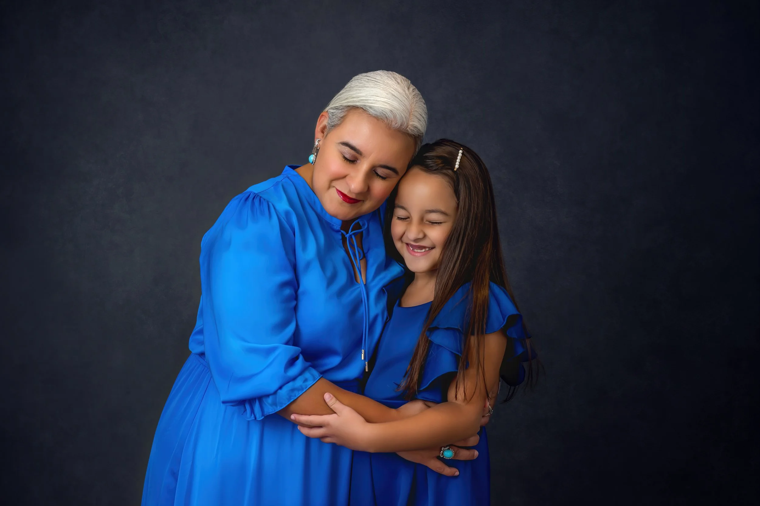 An older woman and a young girl are hugging and smiling with eyes closed, both wearing matching blue dresses, against a dark background.