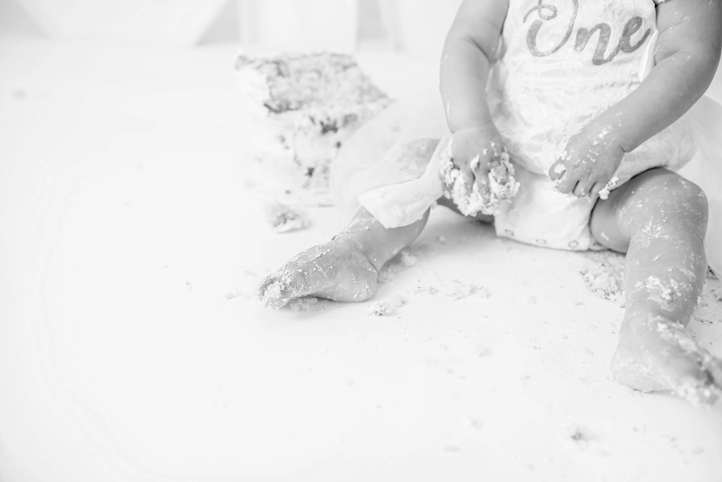 Child sitting on the floor in white clothes, playing with flour and dough, with a piece of cloth and a plate in the background, all covered in flour.