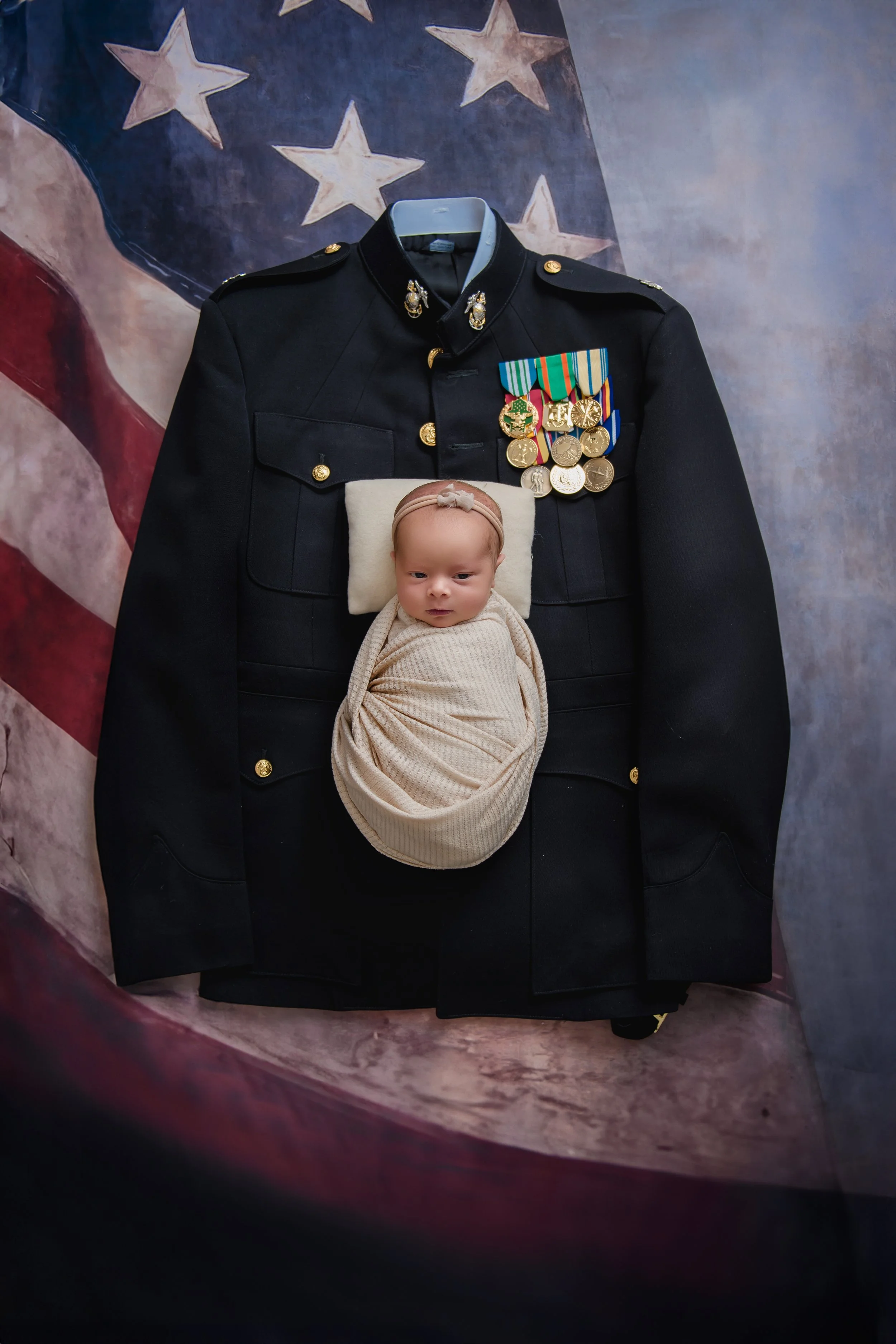 A baby swaddled in a beige blanket, lying on a small pillow, positioned on a US military dress uniform with medals, in front of an American flag backdrop.