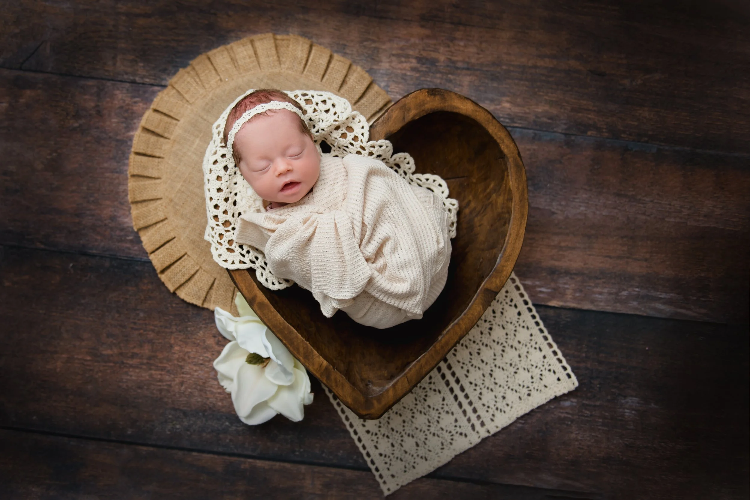 Newborn baby sleeping in heart-shaped wooden bowl, wrapped in beige blanket, with lace doily and white flowers, on wooden floor.