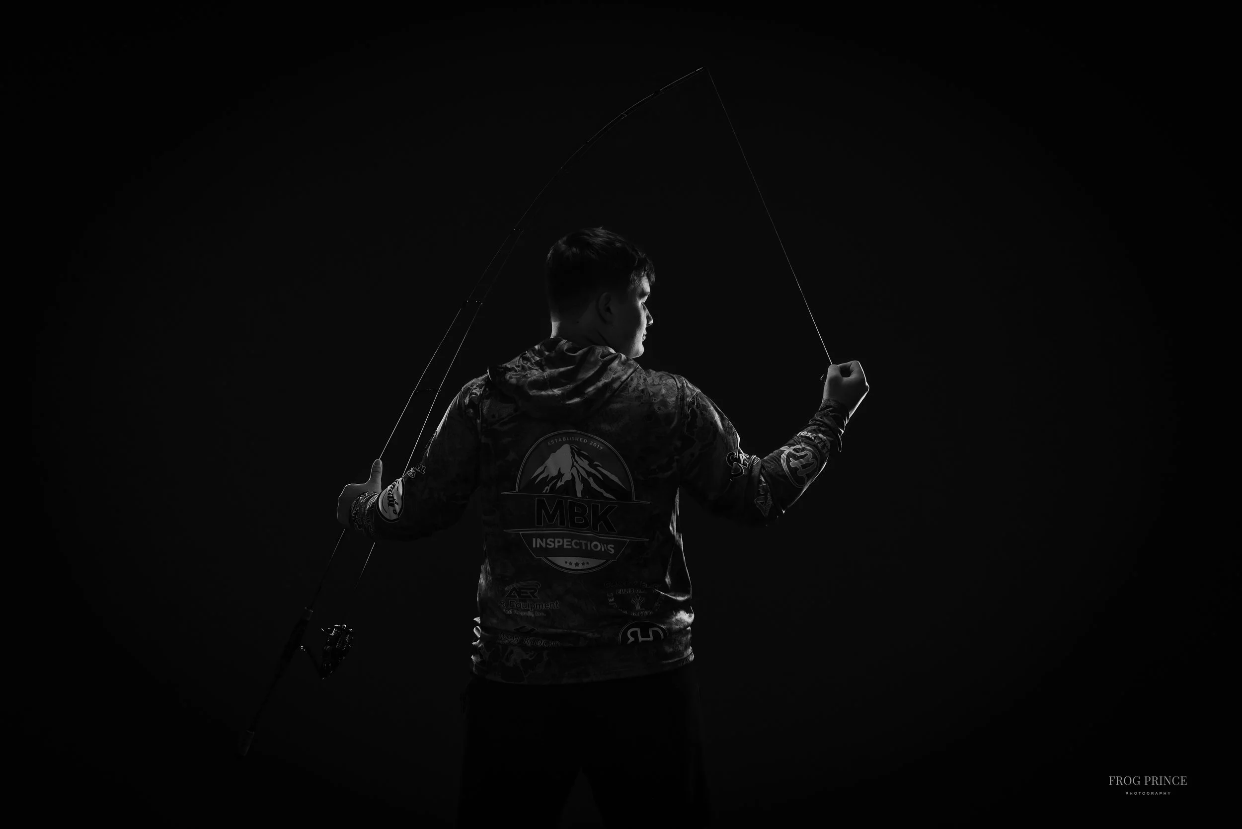 A young man in a hoodie holding a fishing rod against a dark background, facing sideways.