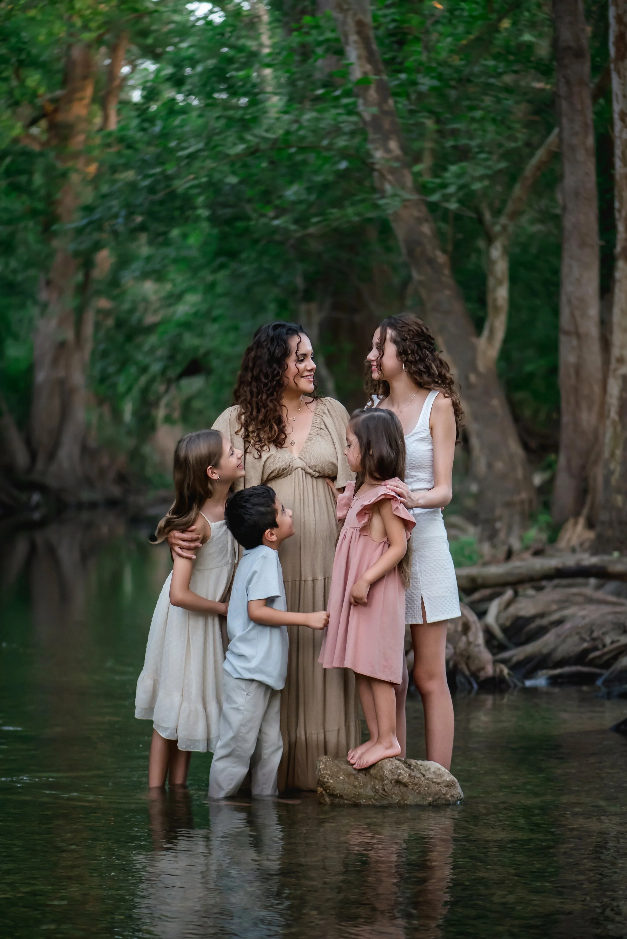 A family of six people standing in a shallow stream in a lush, green forest, looking at each other and smiling.
