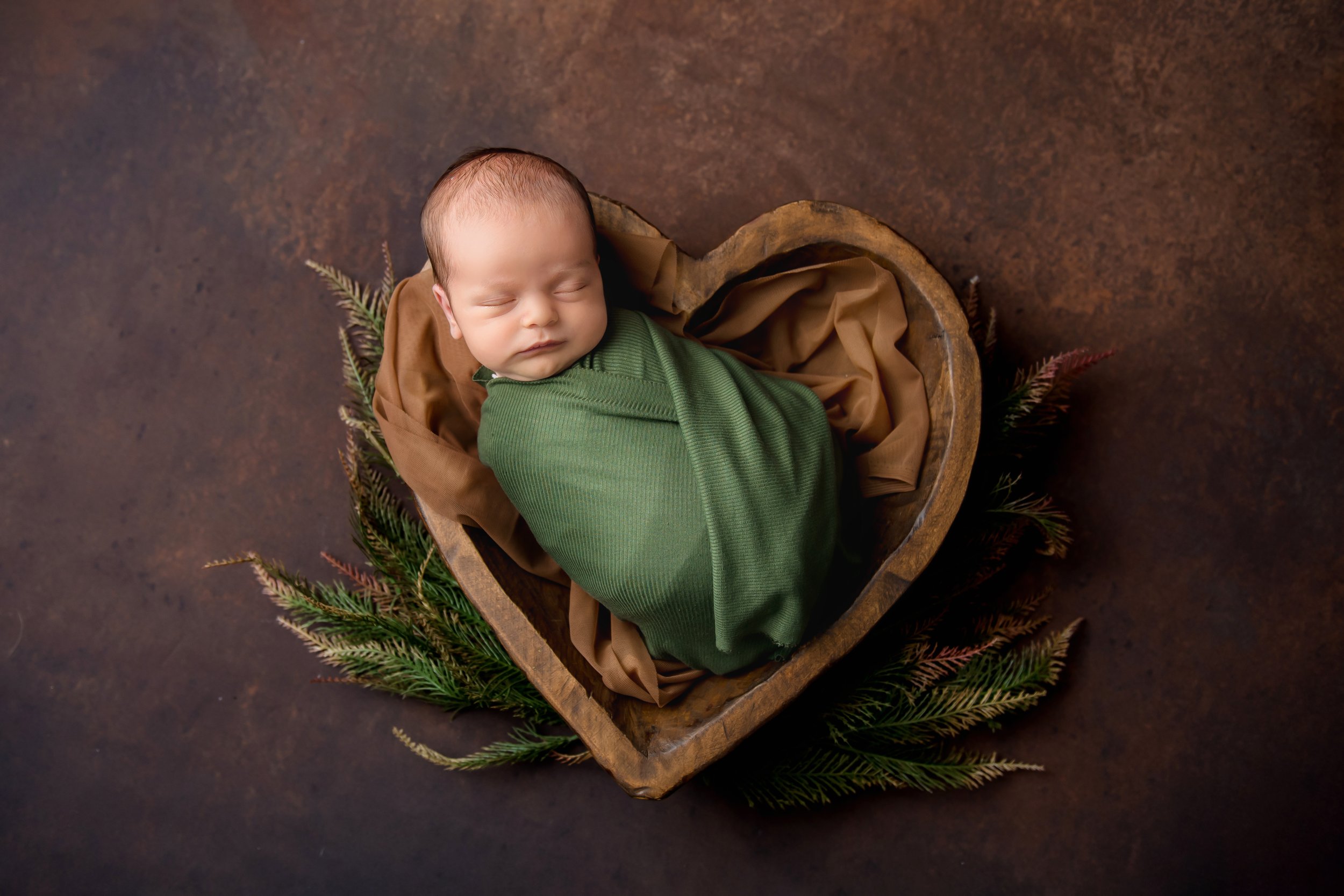 A sleeping baby wrapped in a green blanket, lying inside a heart-shaped wooden frame, with brown fabric lining and green pine branches underneath, on a dark brown background.