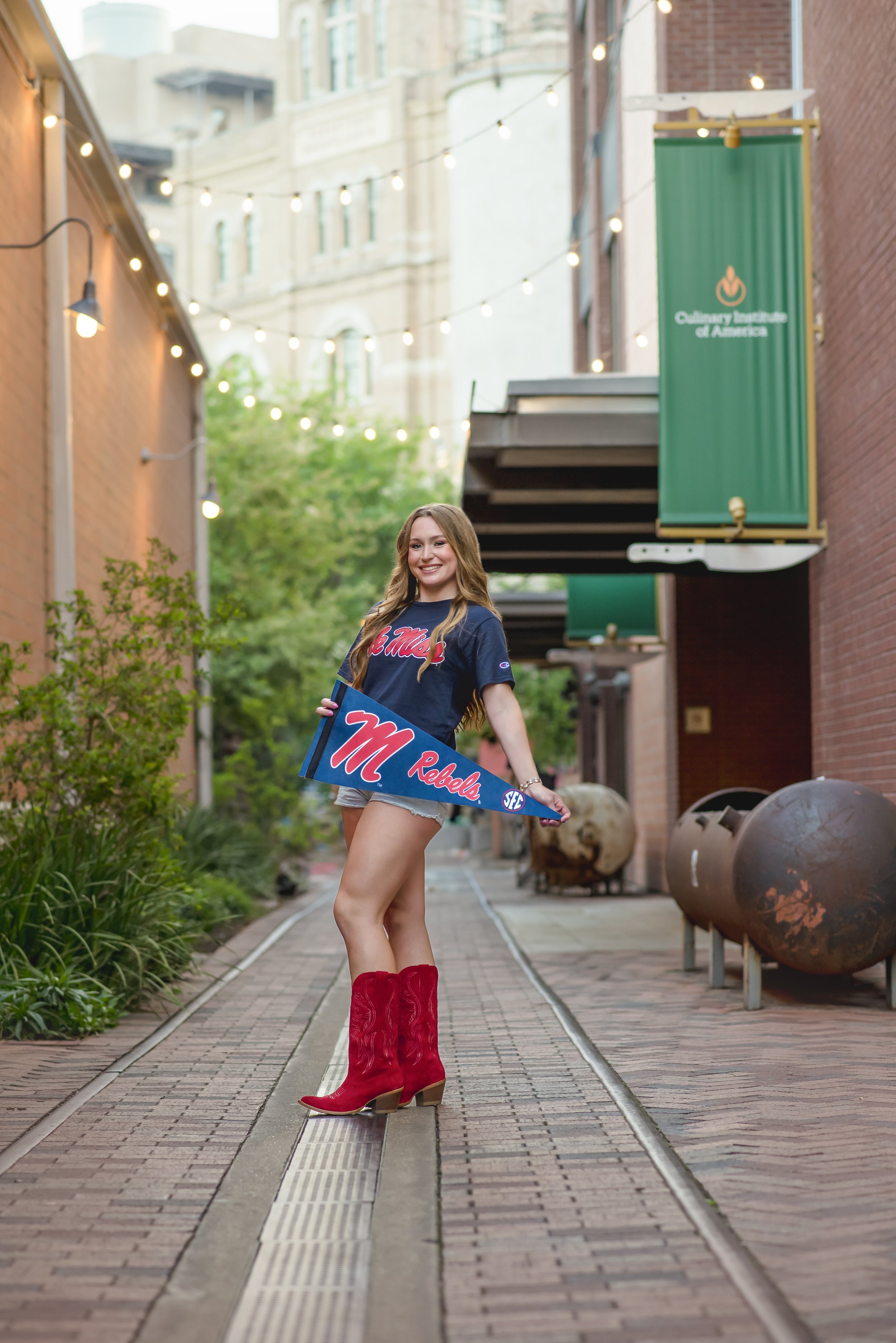 A young woman standing on a brick pathway with railroad tracks, holding a brief flag that reads 'M Rebels.' She is wearing a navy T-shirt, denim shorts, and red cowboy boots, smiling at the camera. The scene is outdoors, lined with string lights and 