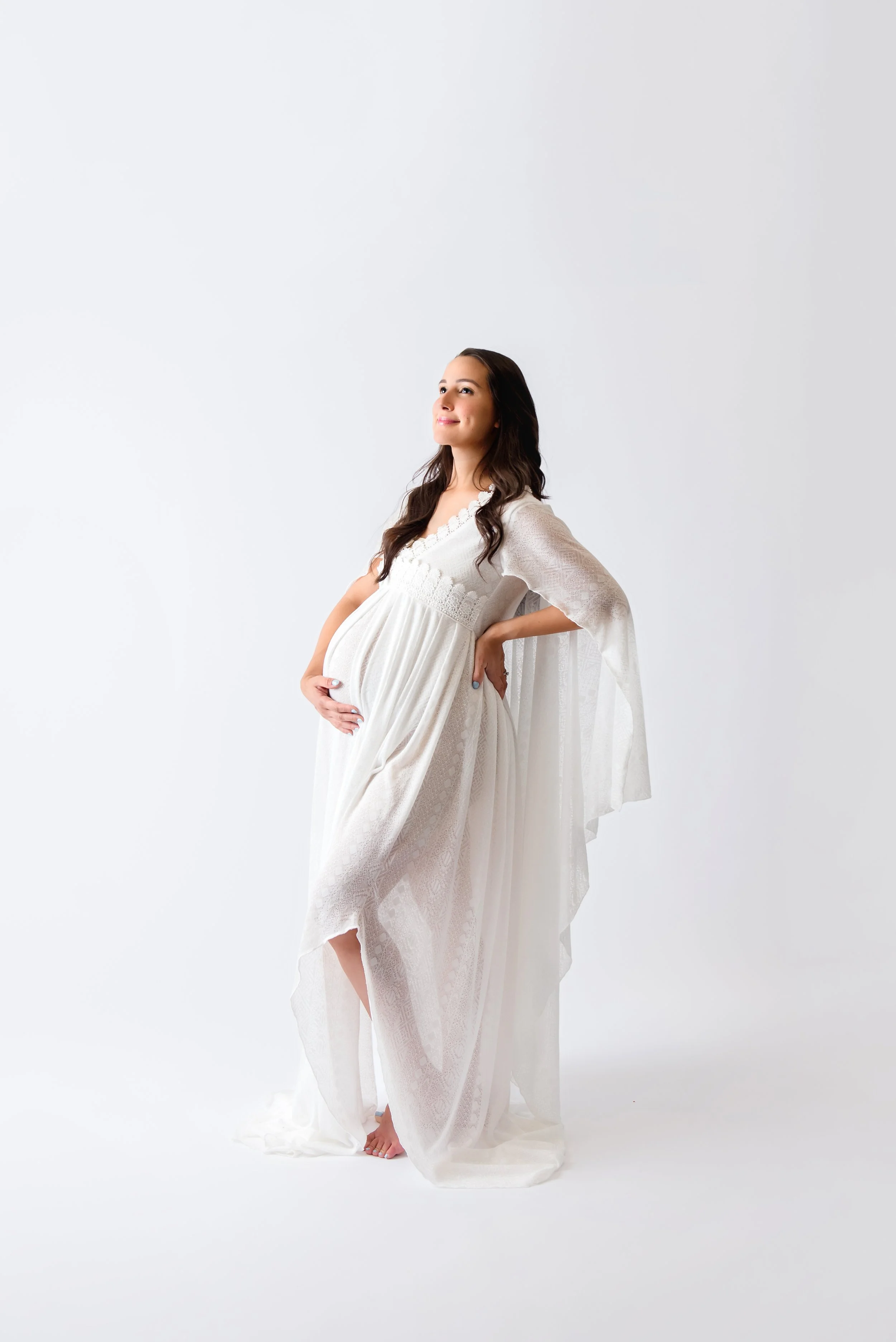 Fine art studio maternity portrait of a mother in a flowing white gown standing against a clean white backdrop, gazing upward with a serene expression.