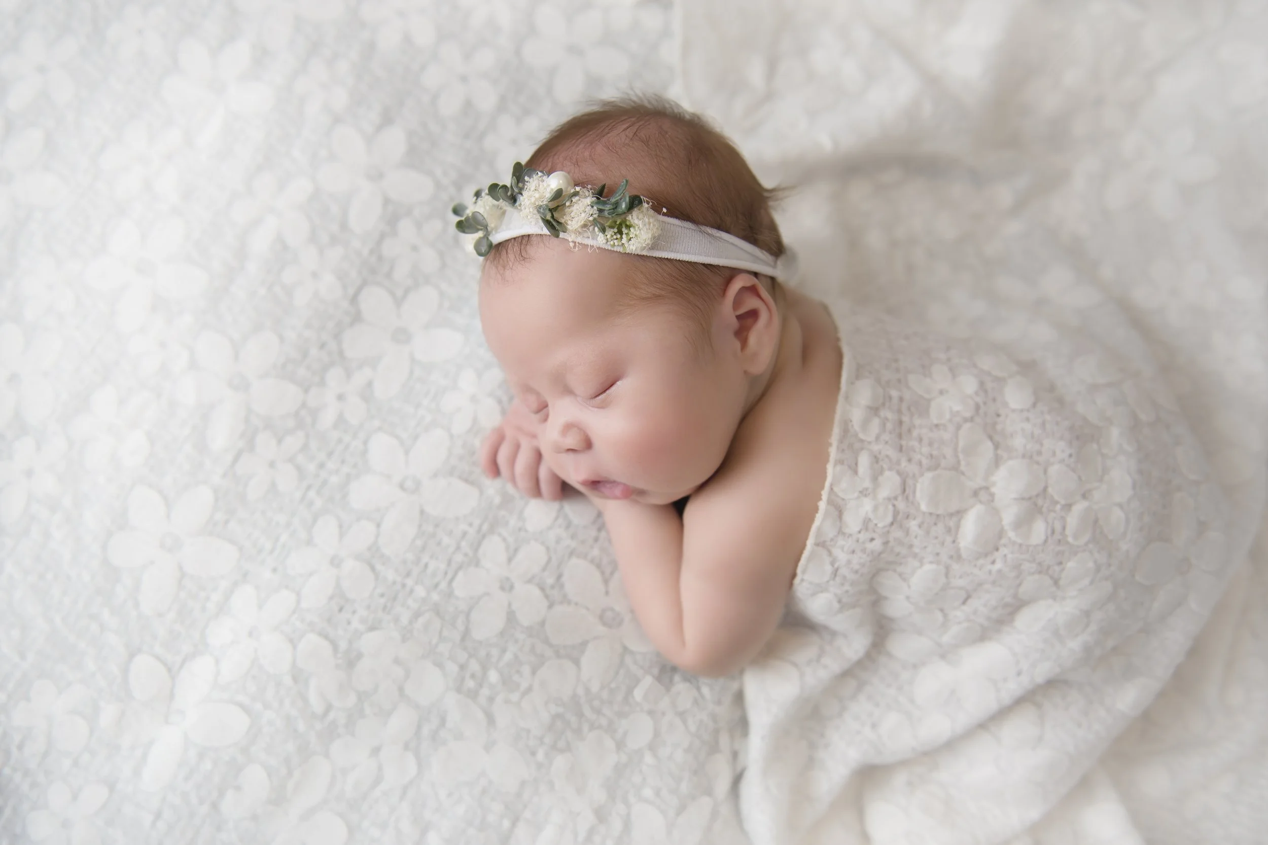 A sleeping newborn baby girl with a floral headband, wrapped in a lace blanket and lying on a textured white surface.