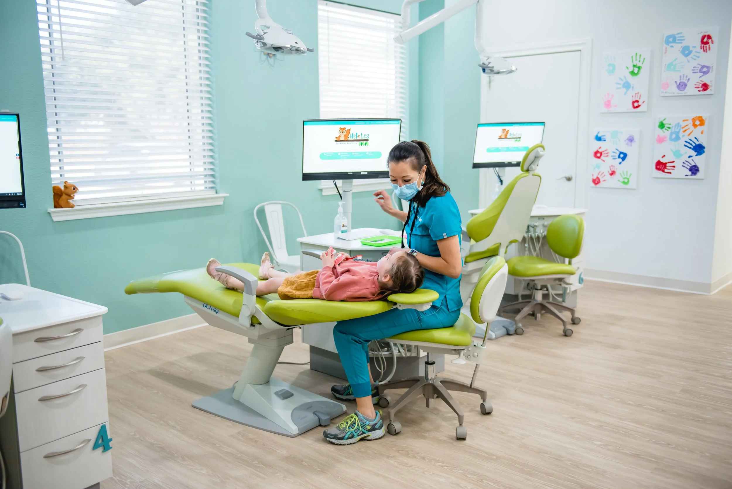 A pediatric dentist or doctor in scrubs and a mask examining a young girl lying on a green dental chair in a colorful dental clinic. The room has light teal walls, white blinds, and children's artwork on the wall.