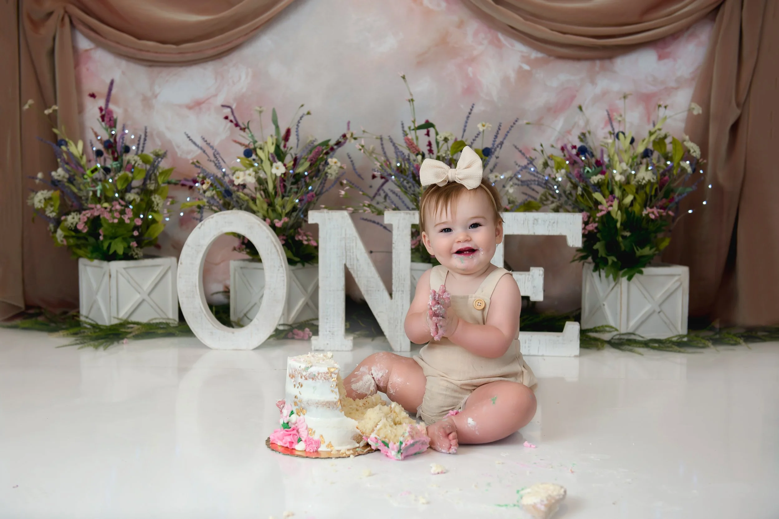 A smiling baby girl with a beige bow in her hair, sitting on the floor with a smashed cake in front of her, surrounded by floral arrangements and large white letters spelling "ONE" for her first birthday celebration.