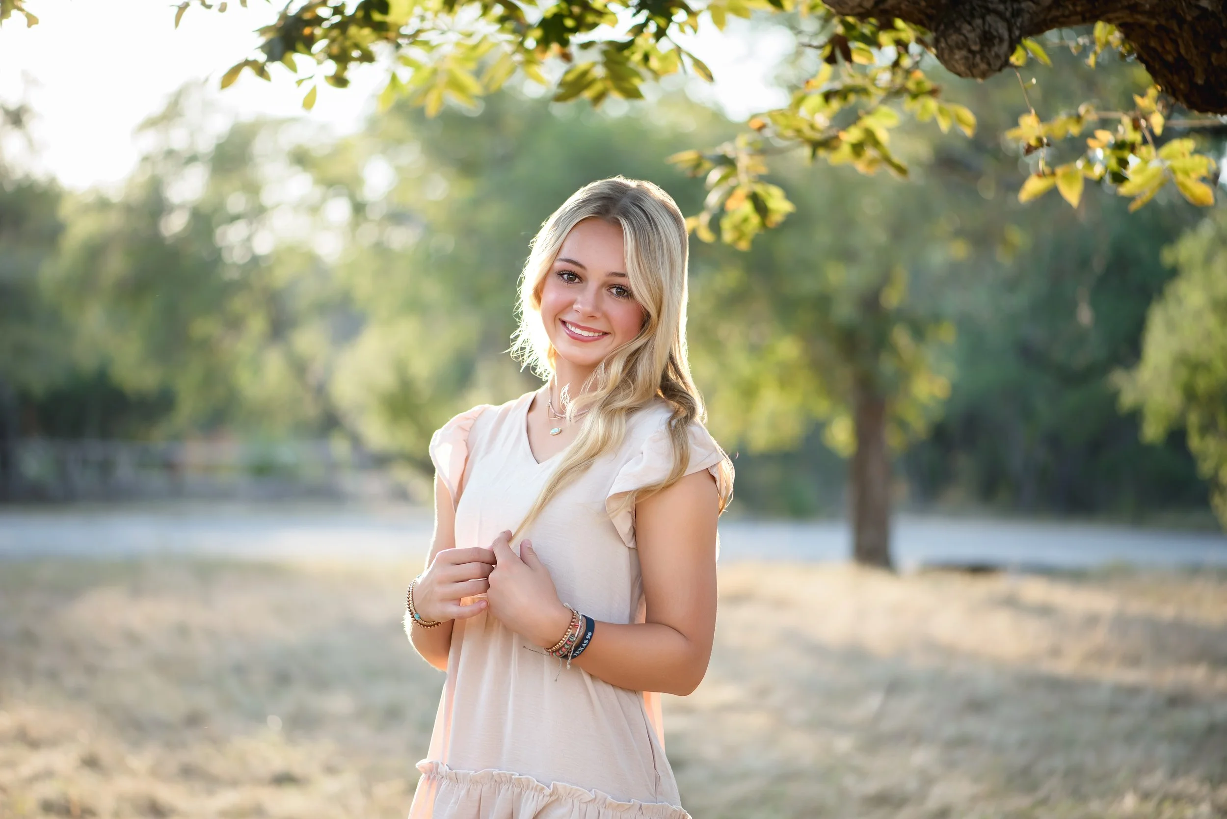 A young woman with long blonde hair, wearing a light-colored dress, standing outdoors under a tree with sunlight in the background. She is smiling and holding a strand of her hair.